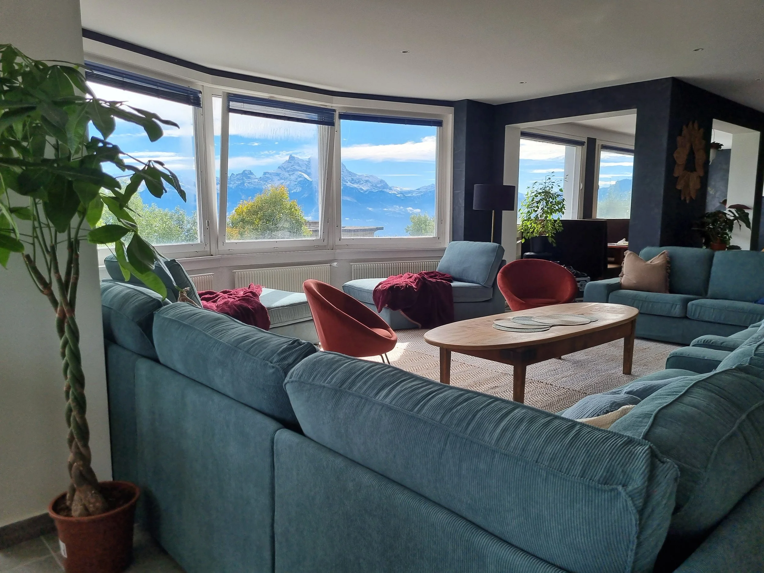 Living room with a large blue sectional sofa, red accent chairs, and a wooden coffee table, featuring large windows with a mountain view.