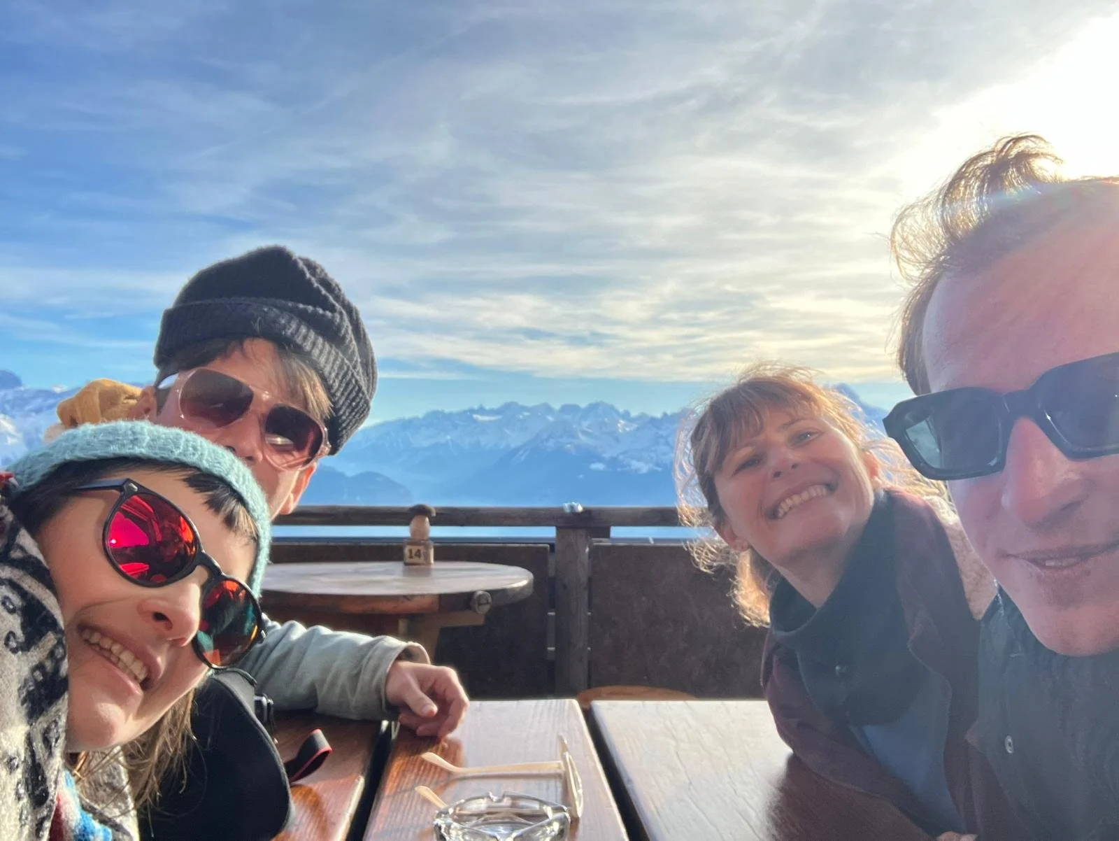 Four people taking a selfie at a mountain overlook during sunset, with snow-capped peaks in the background, all wearing sunglasses and winter clothing.