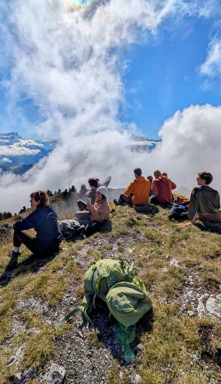 Group of hikers resting on a mountaintop with clouds and mountain peaks in the background.