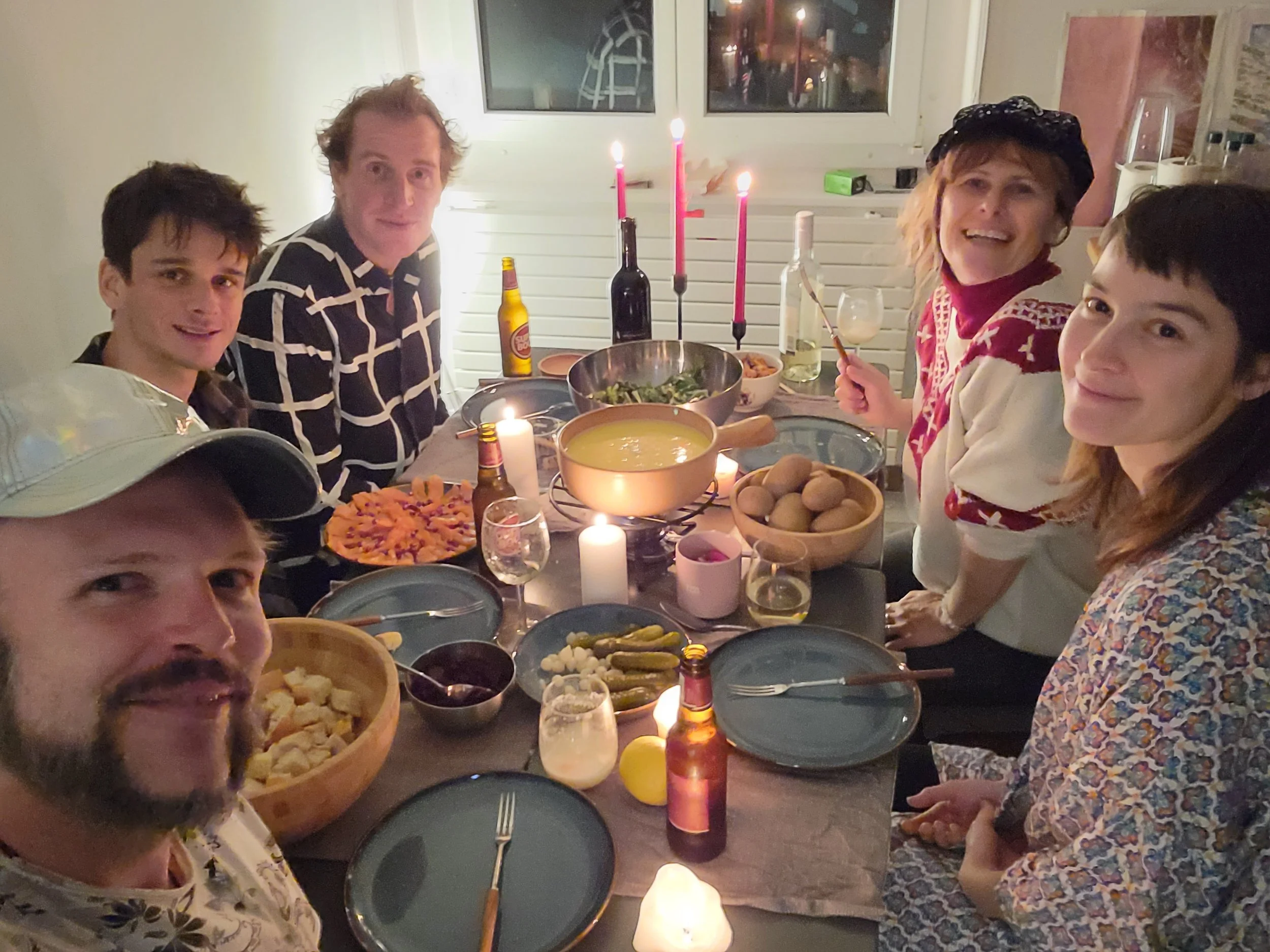 Group of six people gathered around a dinner table decorated with candles, food, and drinks, celebrating together in a cozy indoor setting.