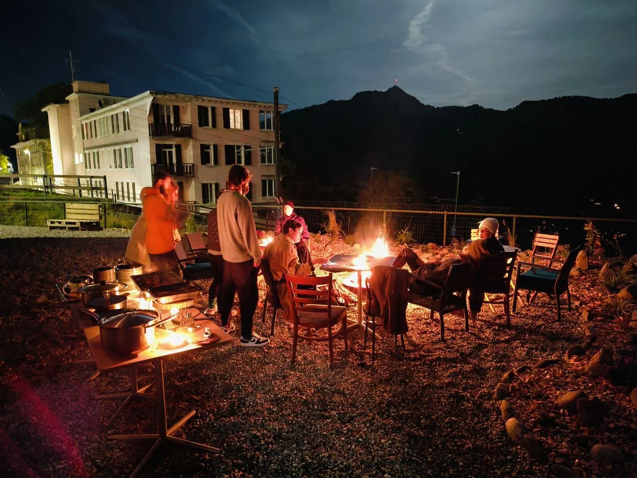 Group of people gathered around a campfire at night outdoors, with a mountainous landscape and a building in the background.