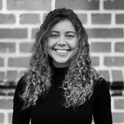 A young woman with curly hair smiling in front of a brick wall.