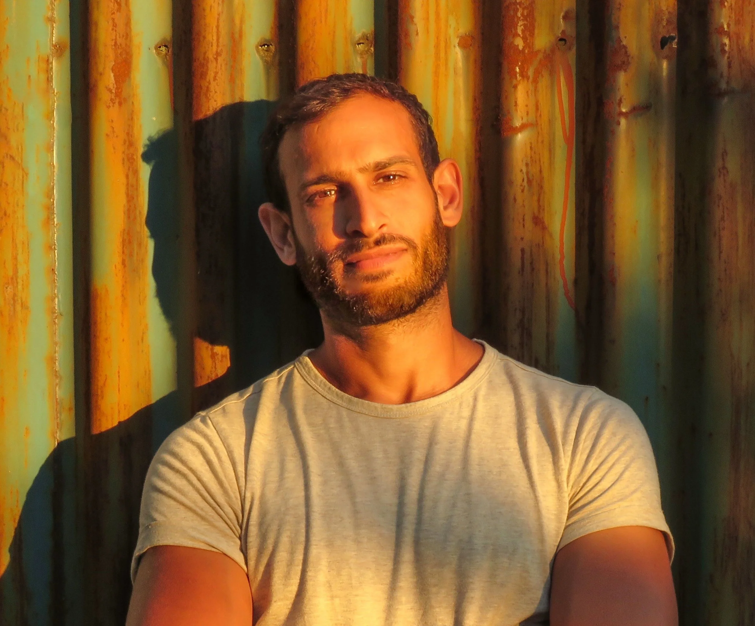 A man with a beard and short dark hair standing outdoors against a rusty corrugated metal wall, illuminated by warm sunlight.
