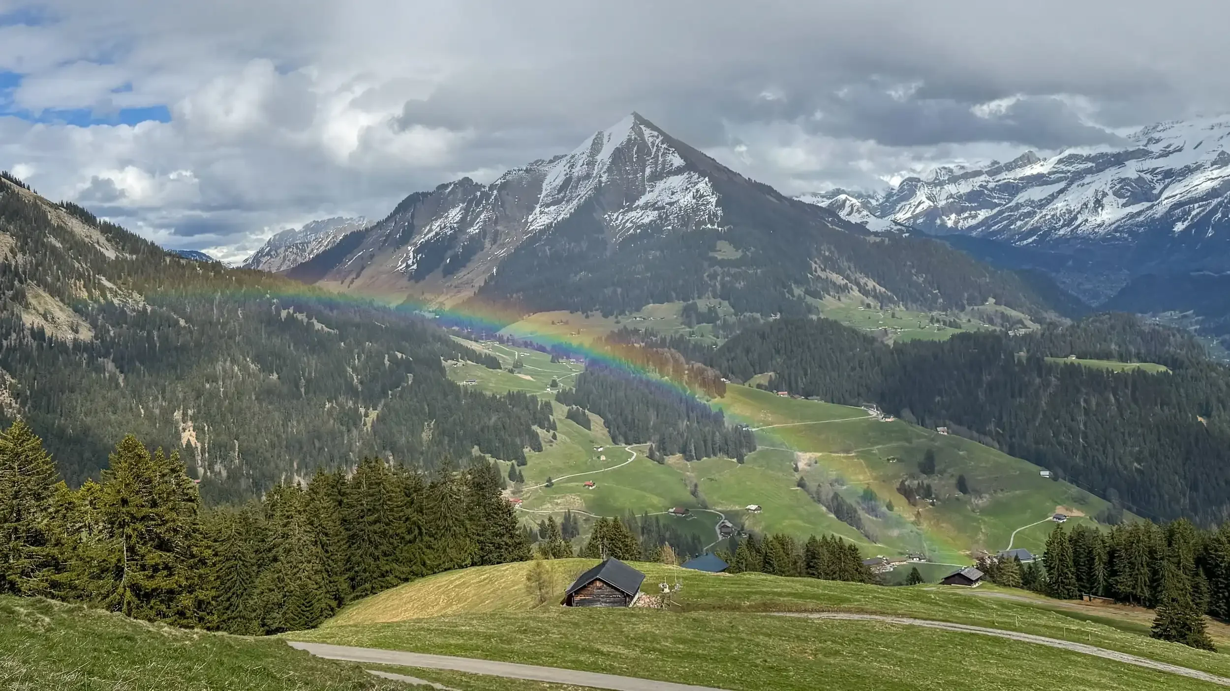 A scenic landscape of green rolling hills and forested mountains with snow-capped peaks under a cloudy sky, featuring a rainbow arching across the valley.