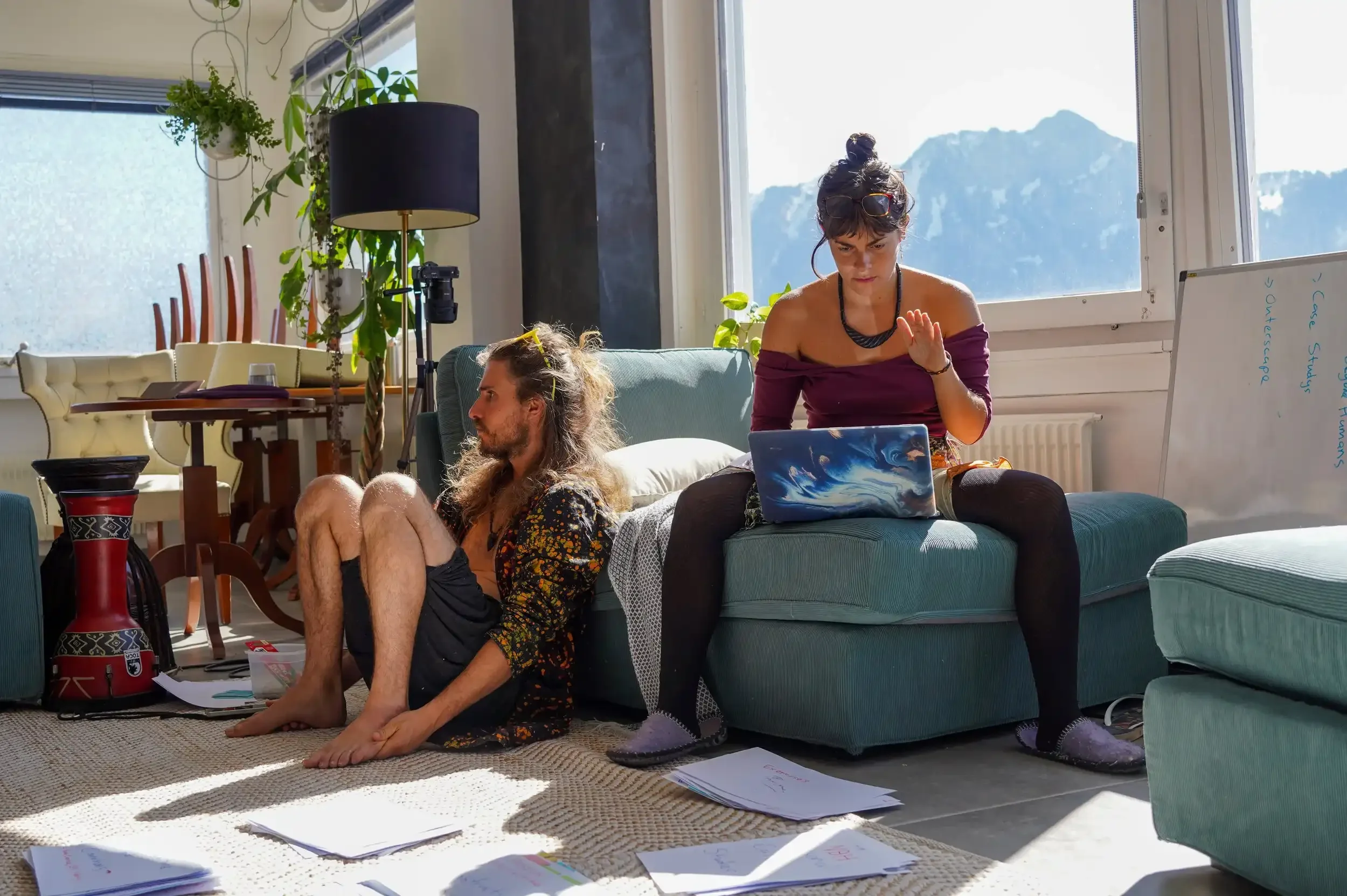 Two young women in a bright living room with large windows, one sitting on the floor and the other on a couch, surrounded by papers and a laptop, with mountains visible outside.