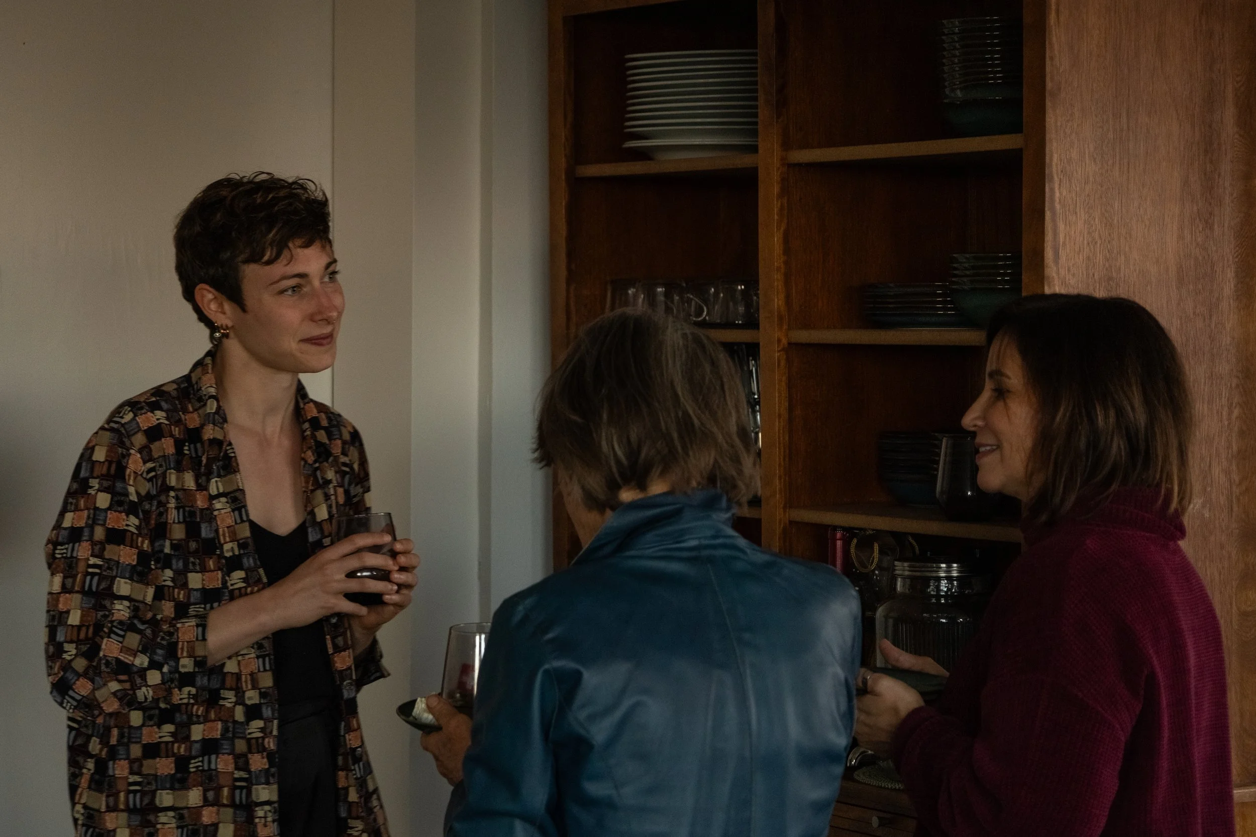 Three women engaged in conversation in a dimly lit room with wooden shelves in the background. One woman on the left is holding a glass of red wine, while the woman on the right is smiling and holding a cup. The woman in the middle has her back to th