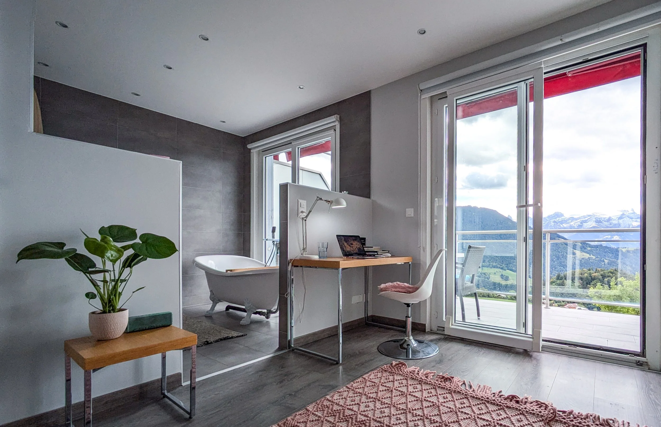 Modern apartment interior with a small workspace, a balcony with scenic mountain views, and a bathroom with a bathtub.