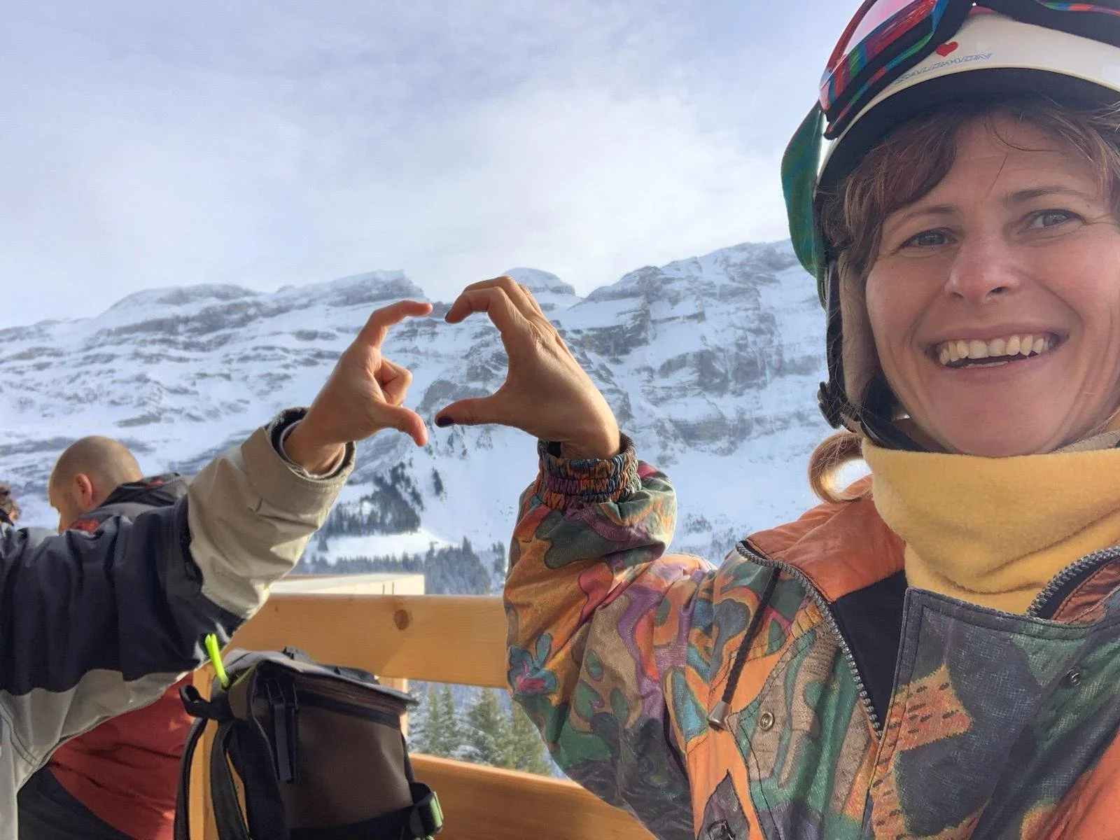 Woman in colorful jacket and helmet smiling and making a heart shape with her hand, snow-covered mountain in the background.