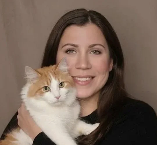 A woman with long dark hair smiling while holding an orange and white cat close to her face.