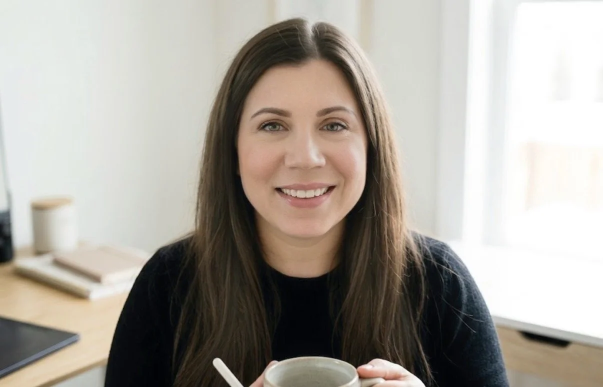 A young woman with long brown hair smiling and holding a mug in a well-lit room.