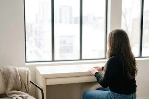 A woman sitting by a large window in a room, gazing outside, with a blanket draped over a chair nearby.