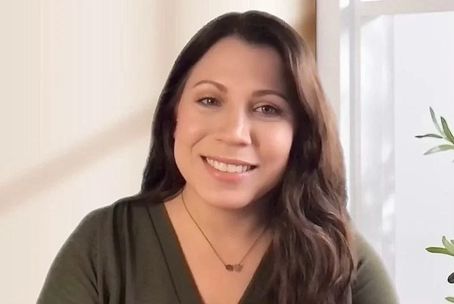 A woman with long brown hair smiling at the camera, wearing a dark green top and a small necklace, sitting indoors near a window with natural light
