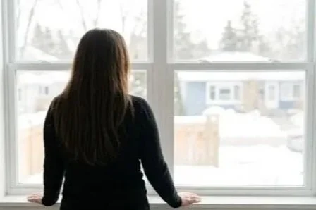 A woman with long brown hair standing in front of a large window, looking outside on a snowy day.