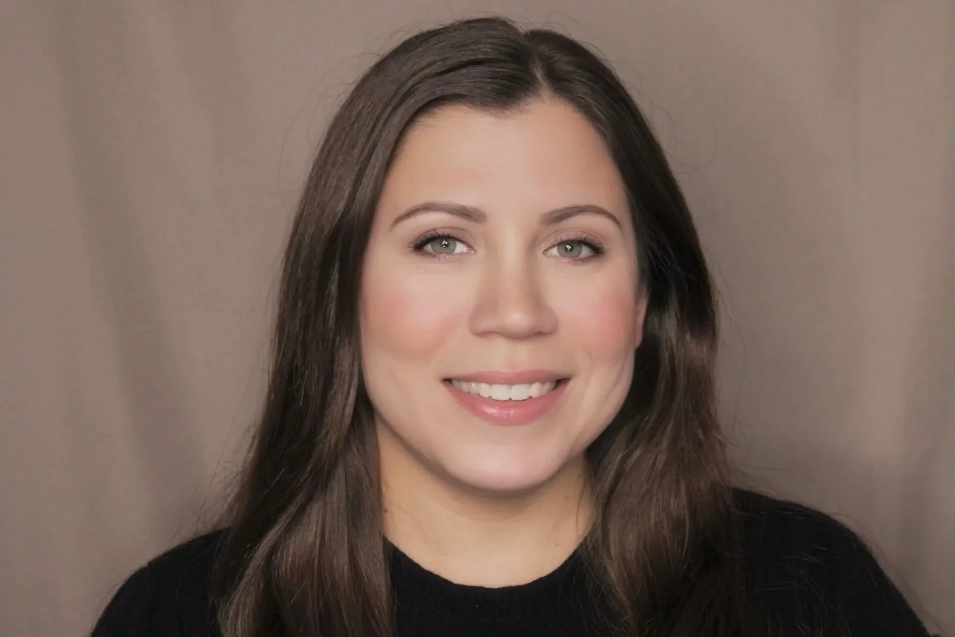 A smiling woman with long brown hair, green eyes, and light skin, wearing a black top, standing against a neutral background.