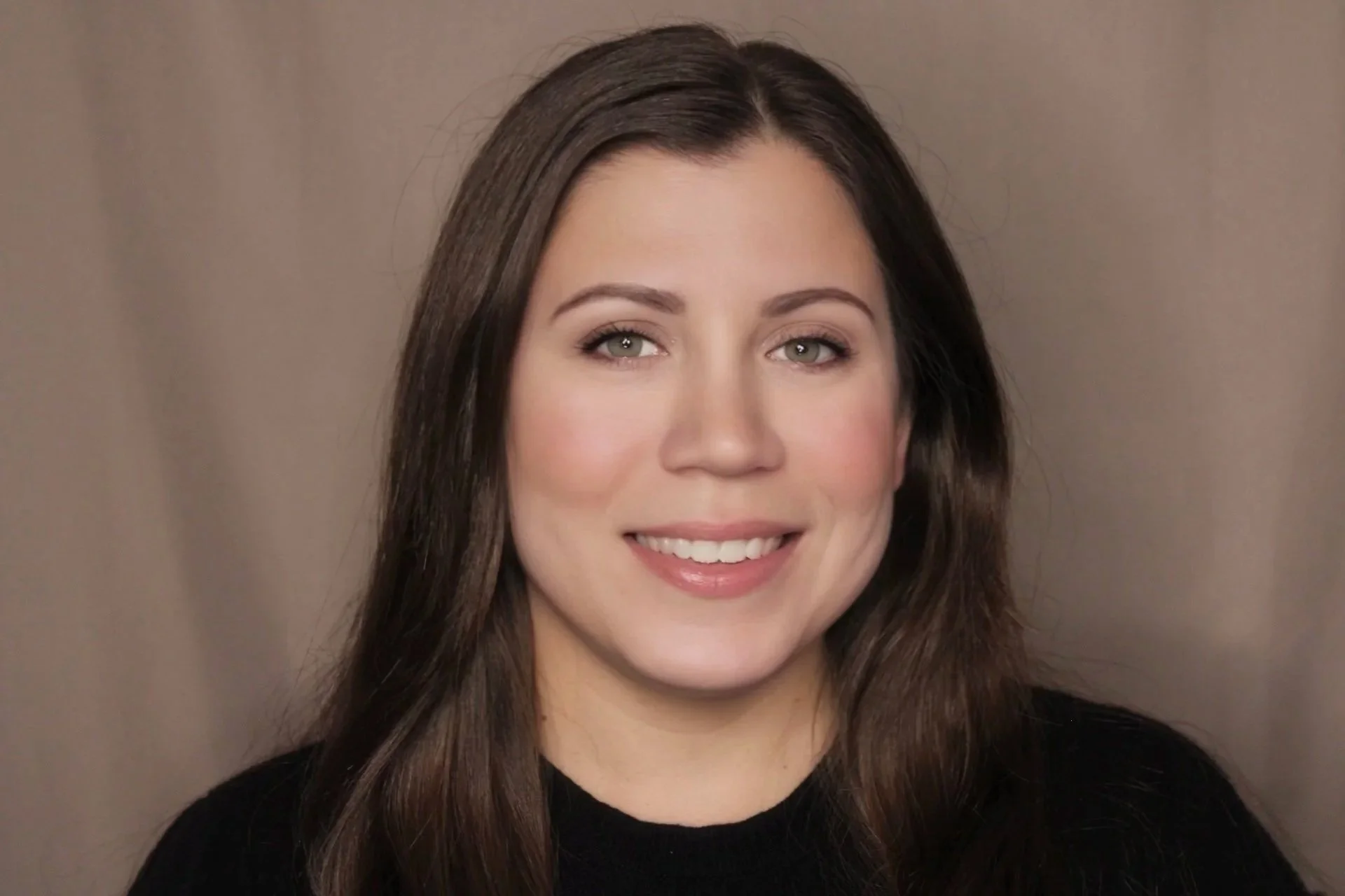 Close-up portrait of a young woman with long brown hair, light skin, and a friendly smile, wearing a black top, against a neutral background.