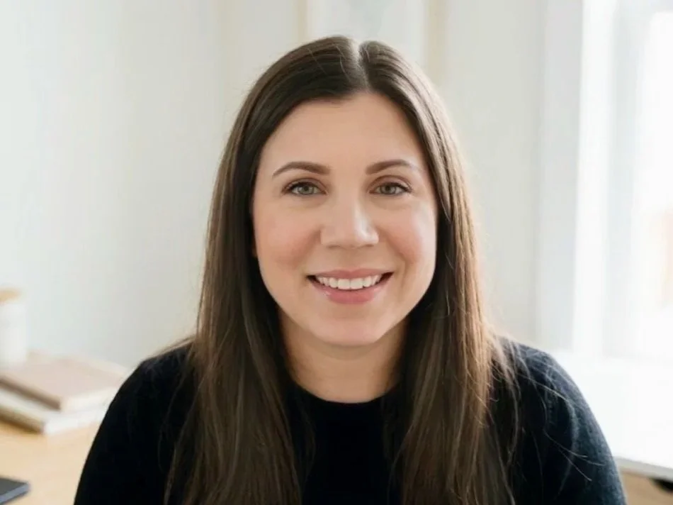 A young woman with long brown hair and a bright smile, wearing a black top, sitting indoors near a window.