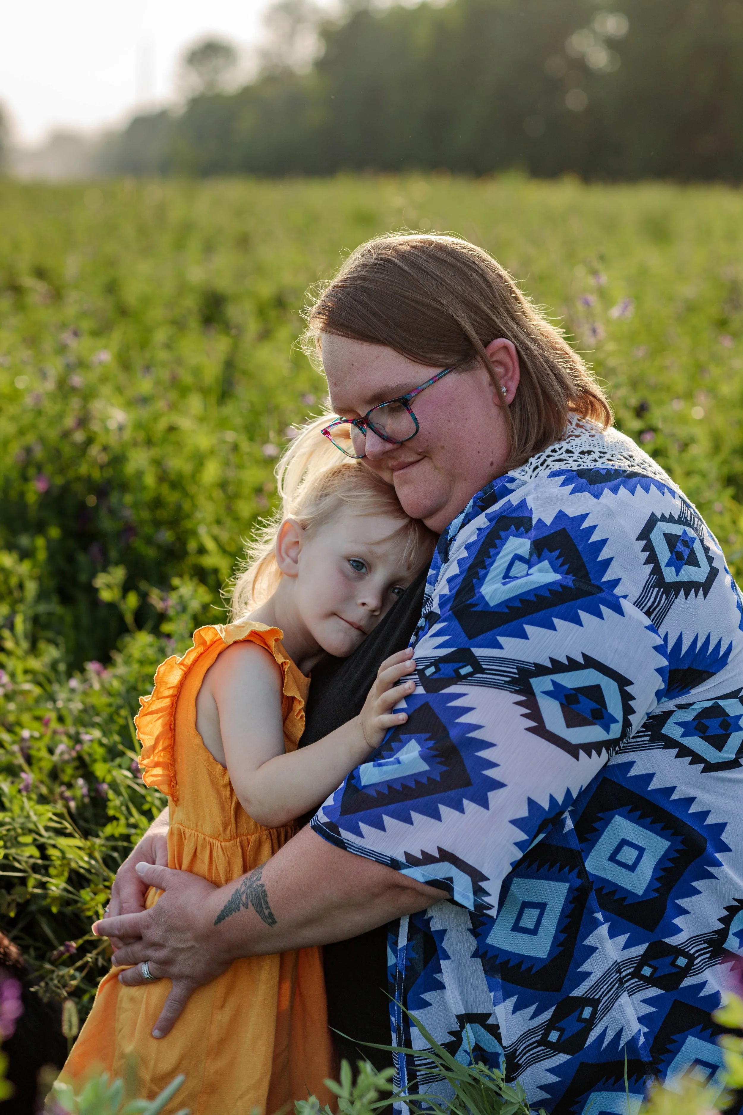 A woman with glasses and shoulder-length hair hugging a young girl with blonde hair and blue eyes in a field of green plants with purple flowers, during sunset.