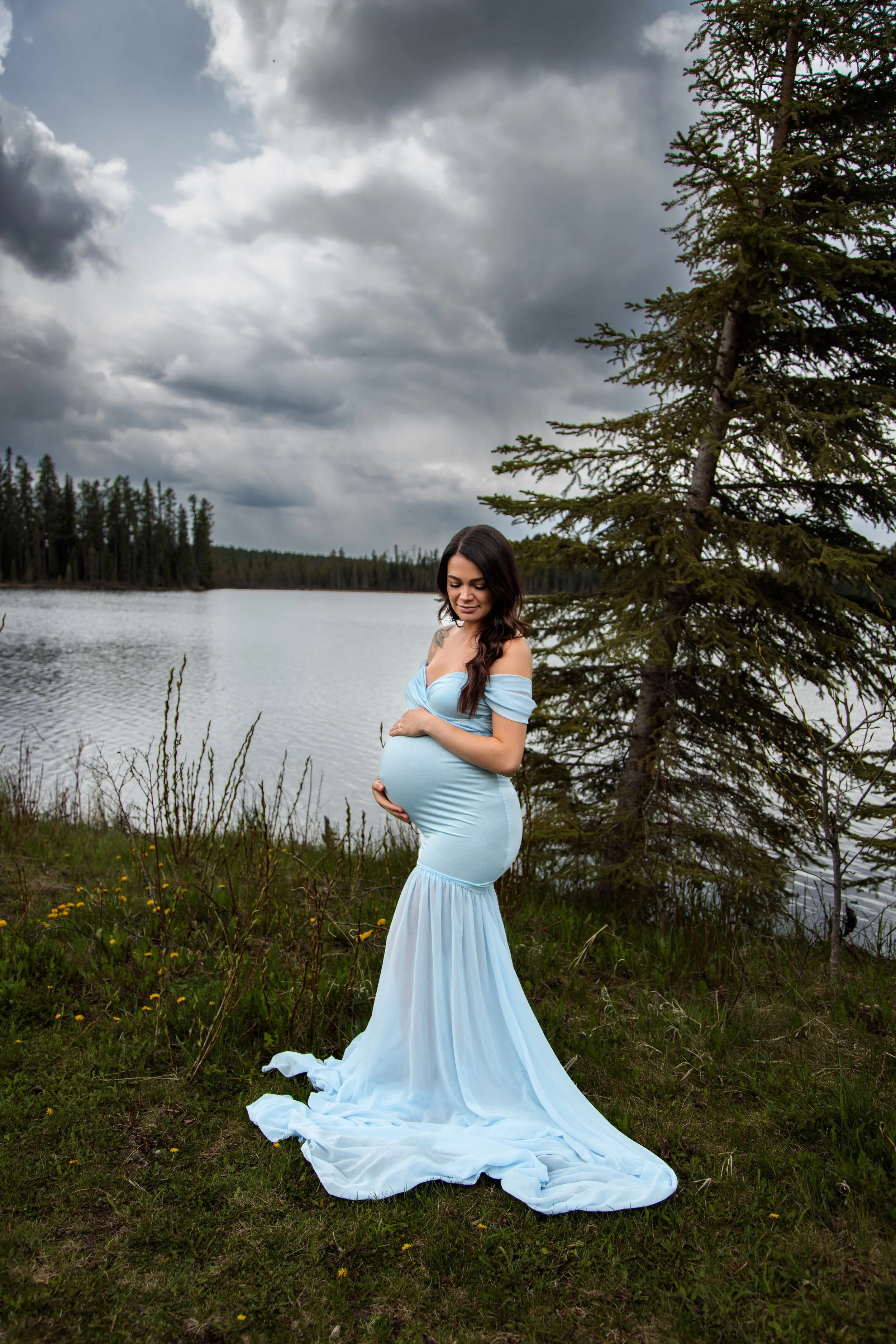 A pregnant woman in a flowing, off-shoulder, light blue gown stands outdoors near a lake with a pine tree on her right. The sky is cloudy and she is gently holding her belly with a serene expression.