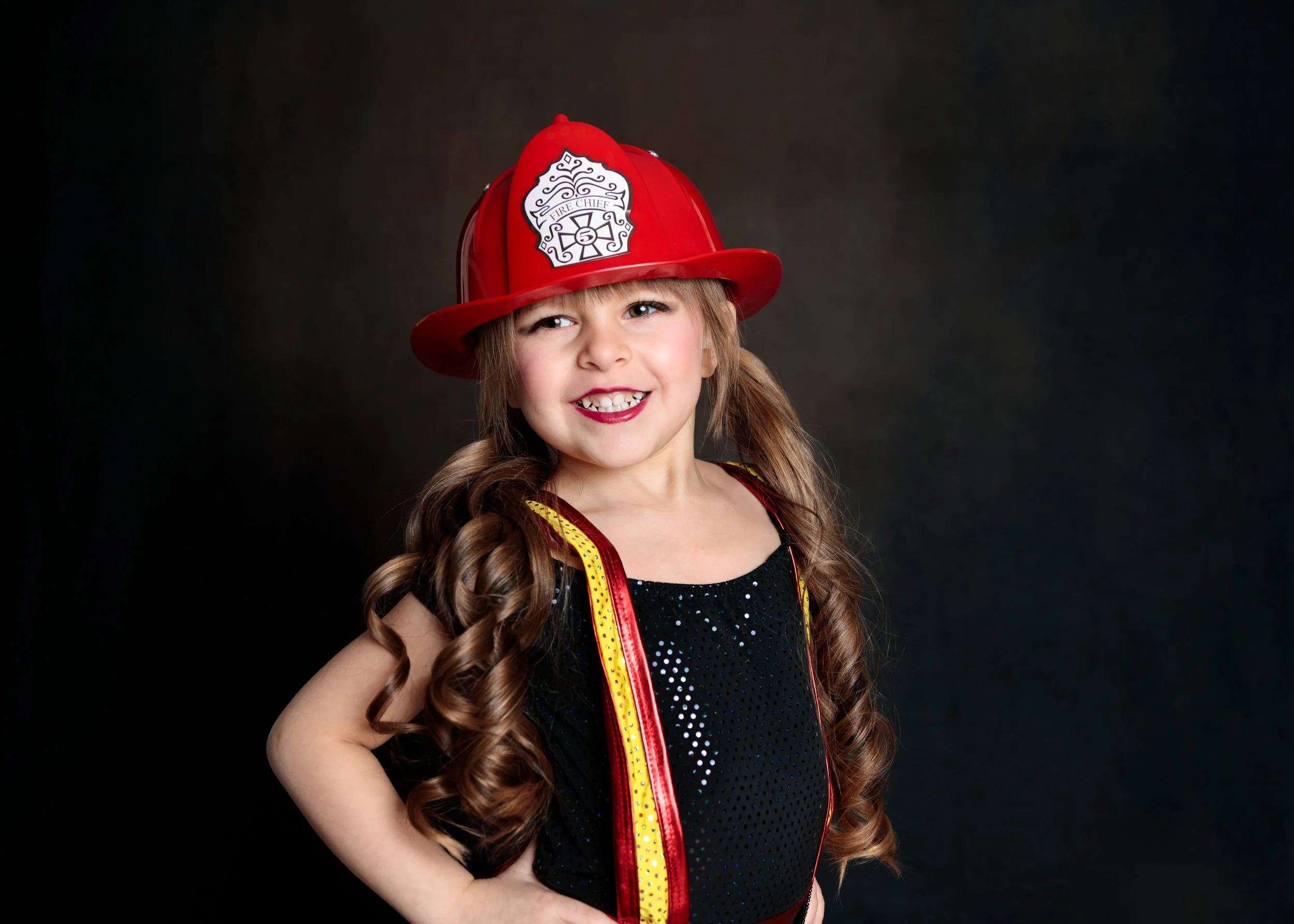 A young girl dressed as a firefighter, wearing a red firefighter hat with a badge reading 'Fire Chief' and a black dress with sparkles, smiling and standing against a dark background.