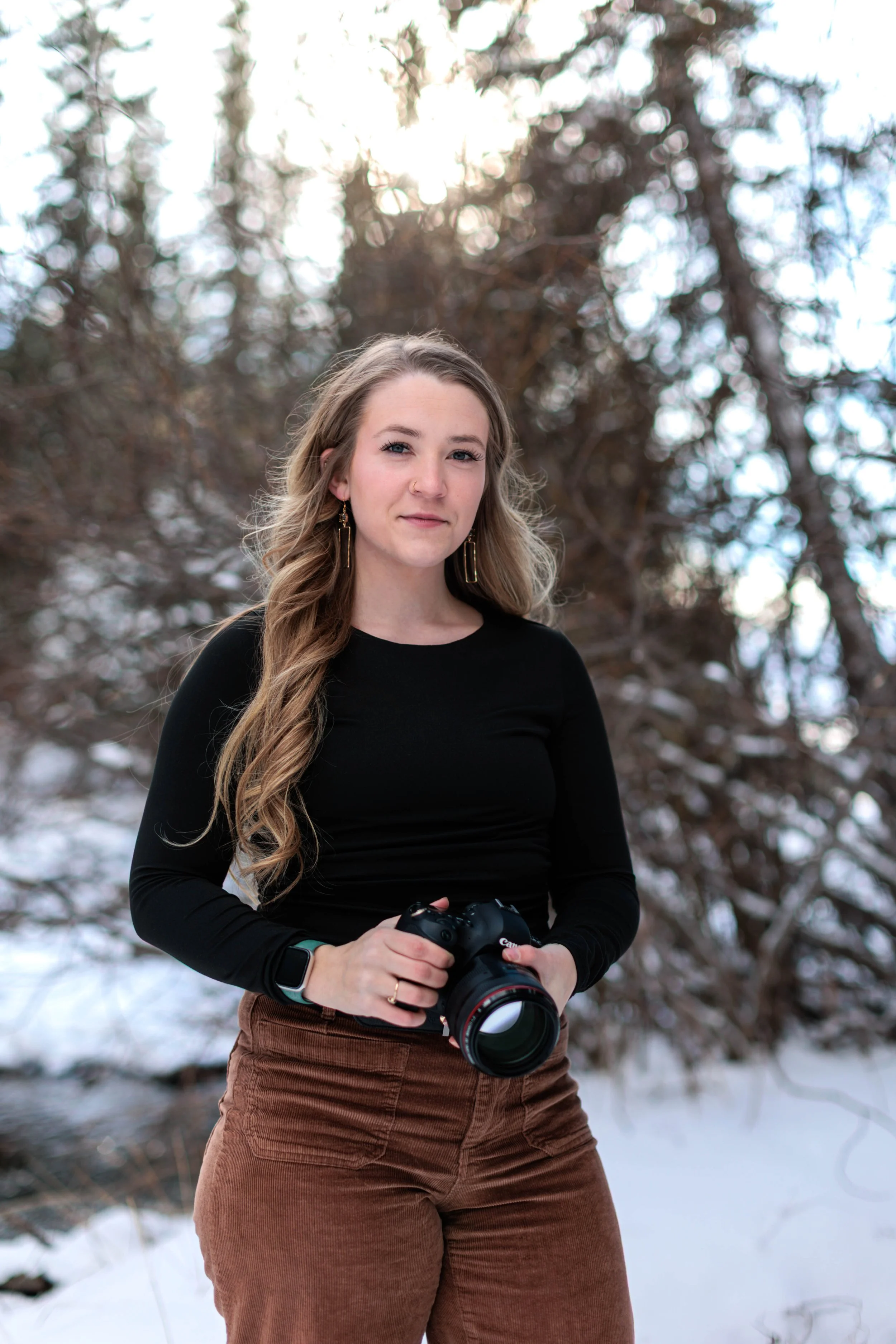A woman with long wavy hair holding a camera outdoors in a snowy, wooded area.
