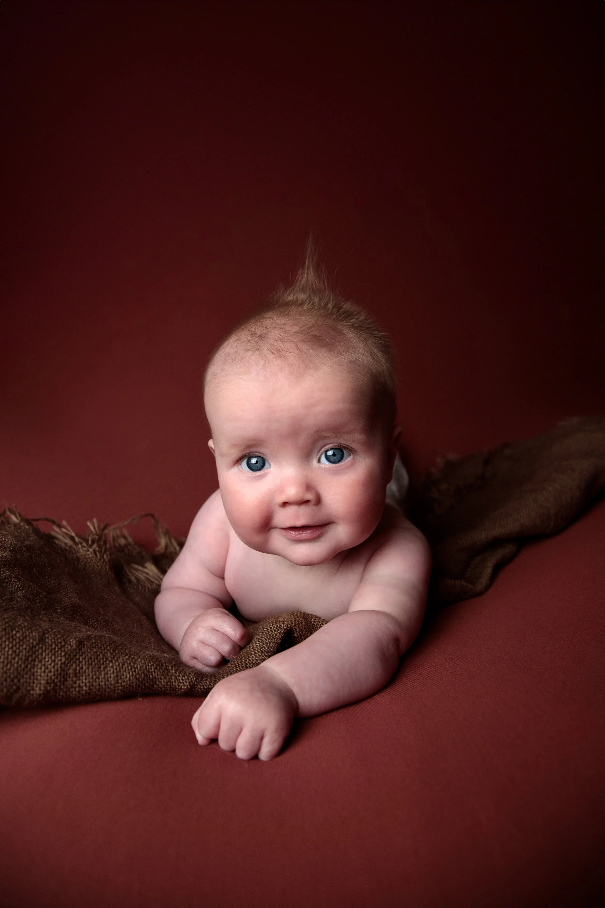 A baby with blue eyes and a tuft of light hair, lying on a brown blanket and propped up on their arms against a red background.