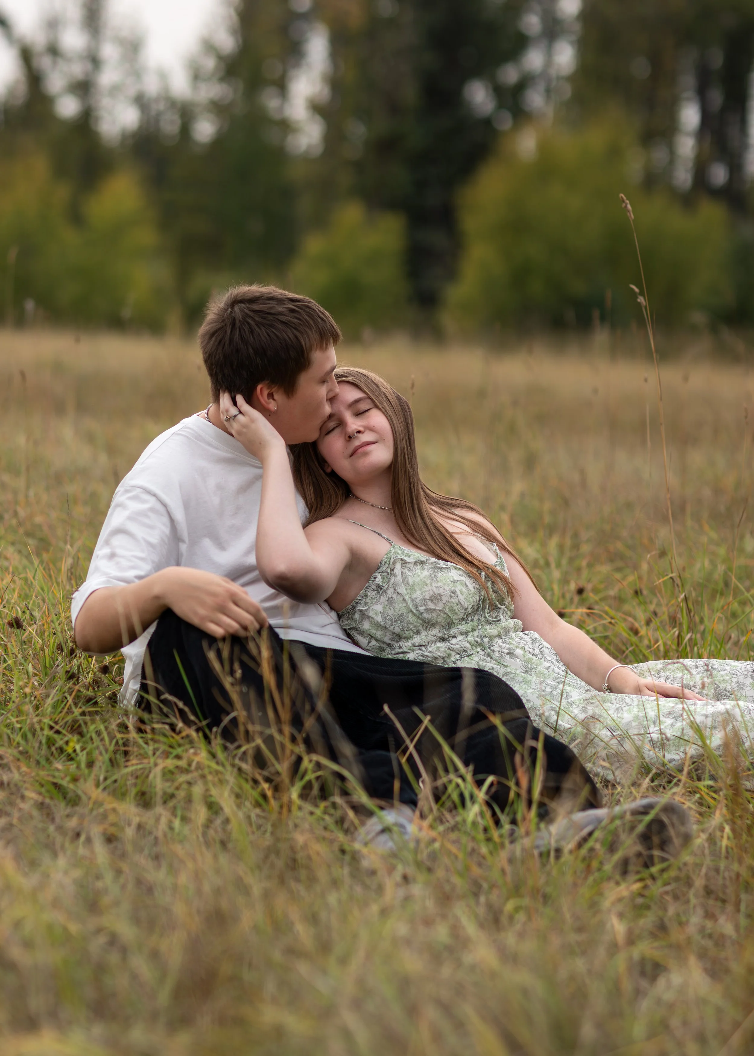A young couple sitting in a grassy field with trees in the background. The man is kissing the woman on her forehead while she relaxes with her eyes closed, holding her hair back with one hand.