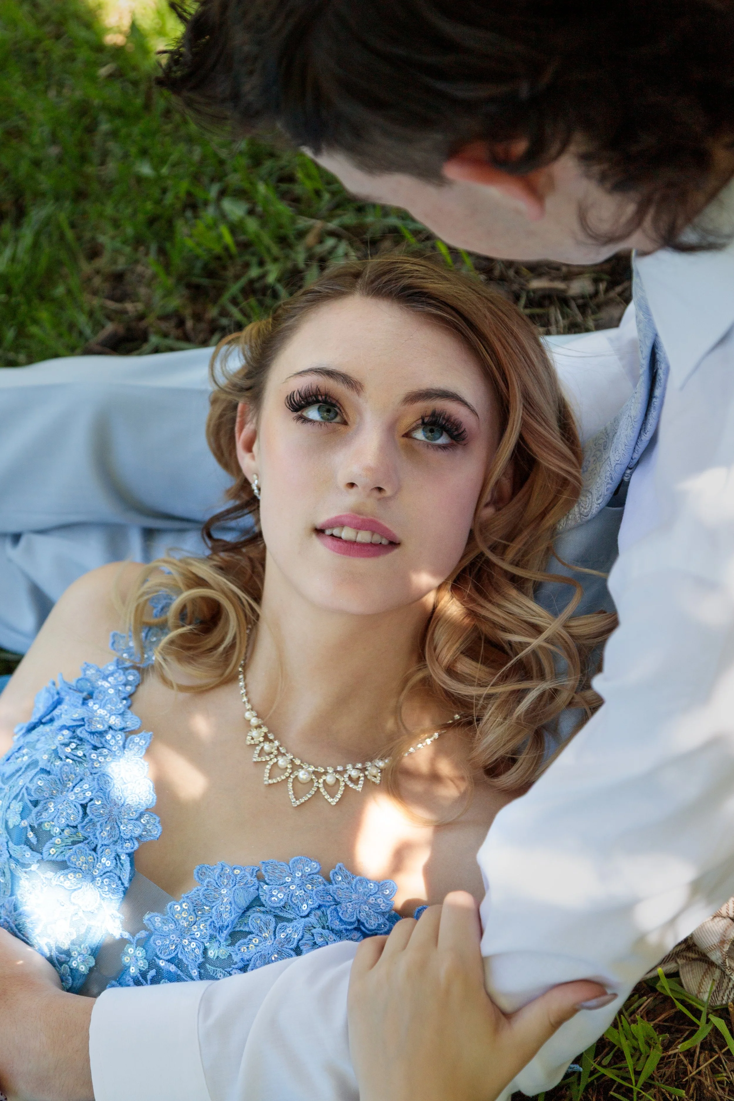 A couple lying on grass, gazing at each other, with the woman wearing a blue dress and pearl jewelry, and the man in a white shirt.
