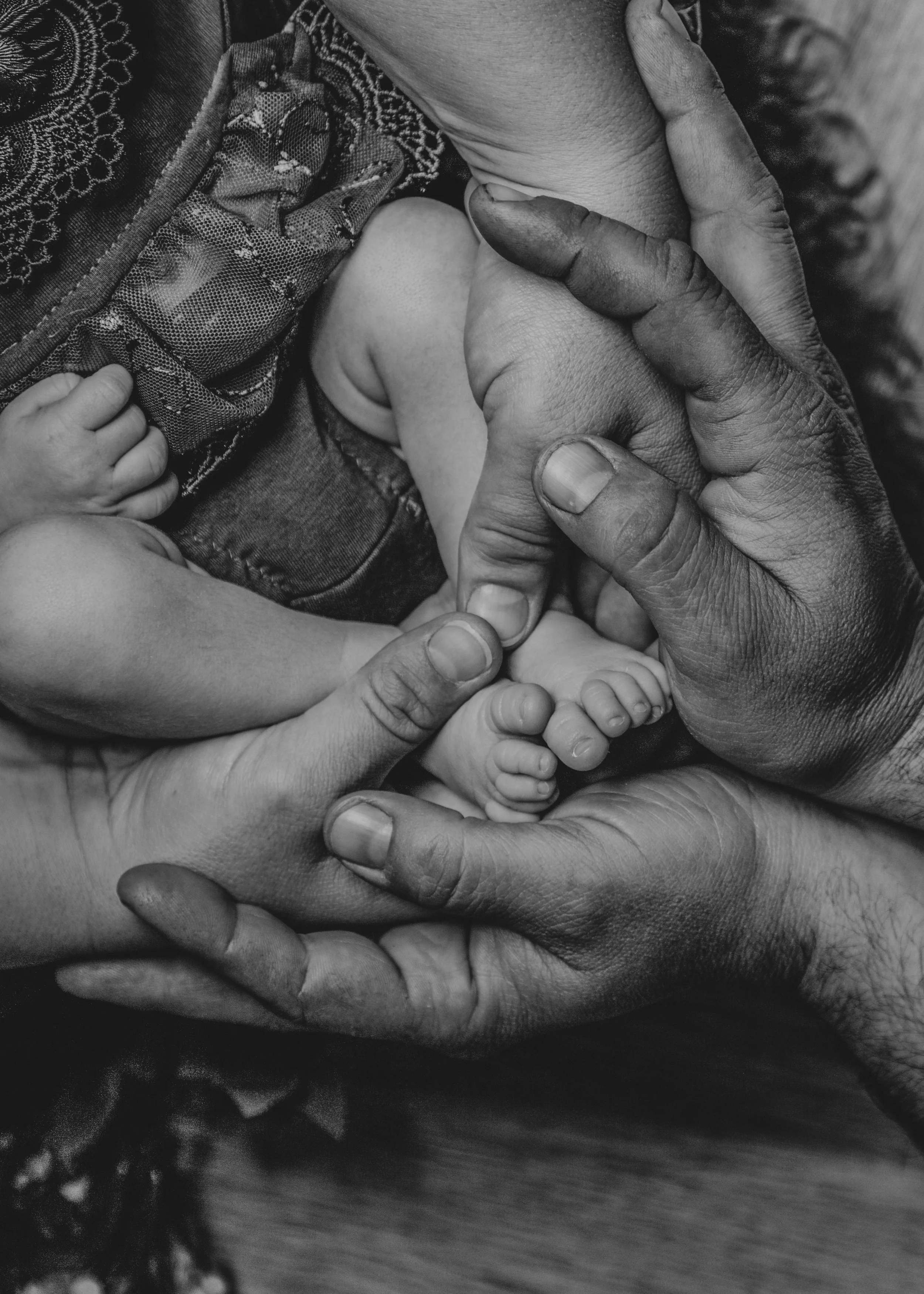 A close-up black and white photograph of multiple hands and fingers gently holding a small baby’s foot, with some wearing patterned clothing.