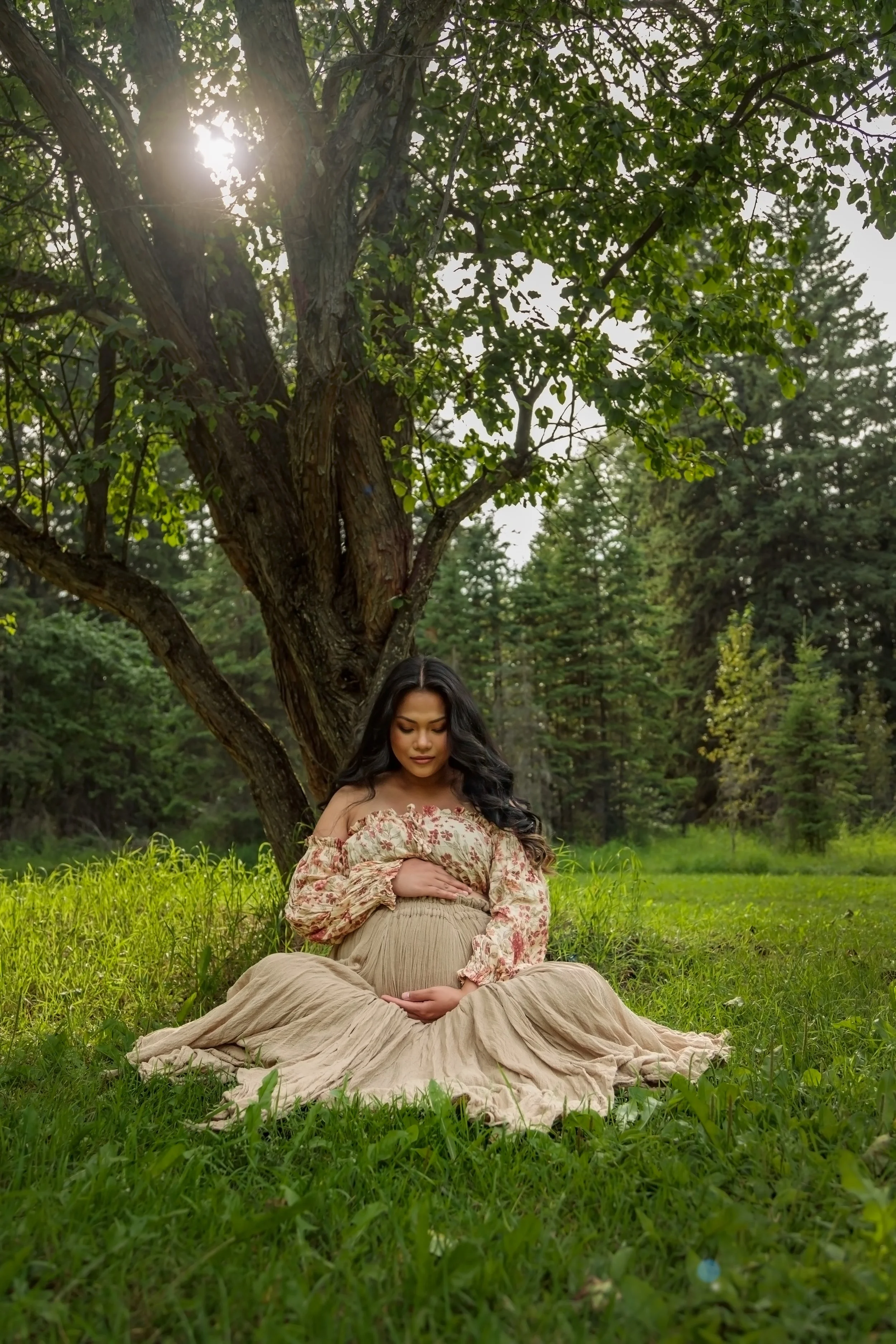 A pregnant woman sitting cross-legged on green grass under a large tree in a natural outdoor setting. She is cradling her belly with both hands and looking down with her eyes closed. The woman is wearing a floral off-shoulder blouse and a long beige 