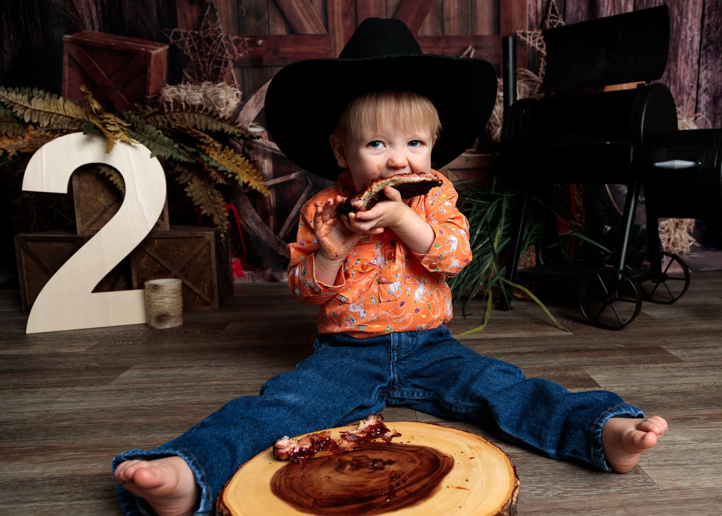 A young child with blonde hair, wearing a black hat and orange shirt with animal patterns, sitting on a wooden floor, eating pizza. A person, possibly an adult, is lying on the floor with legs spread apart, with a wooden serving platter with pizza sl