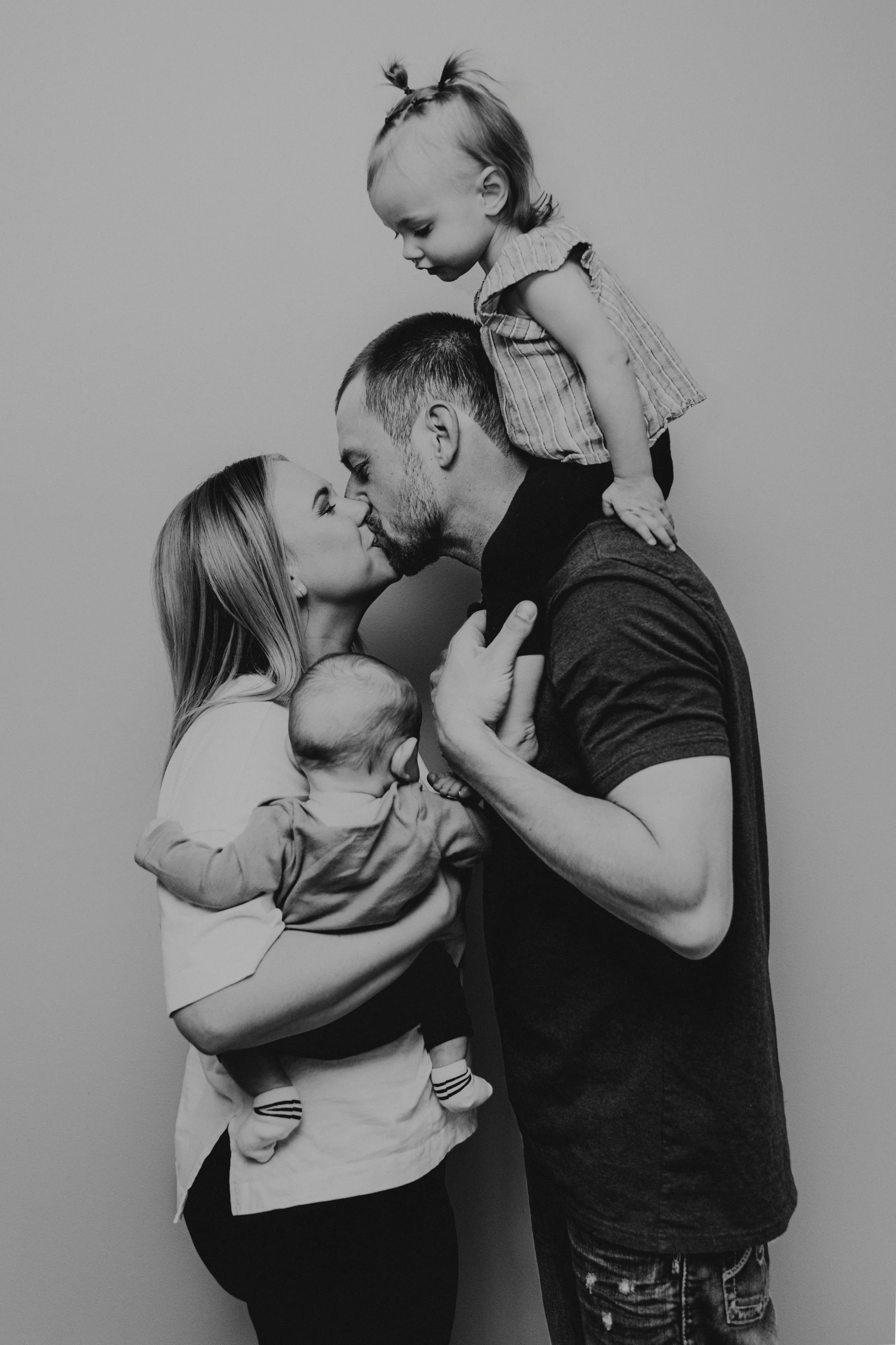 A black and white photo of a family of four, with a woman and a man kissing, a young girl sitting on the man's shoulders leaning toward him, and a small child in the woman's arms, all against a plain background.