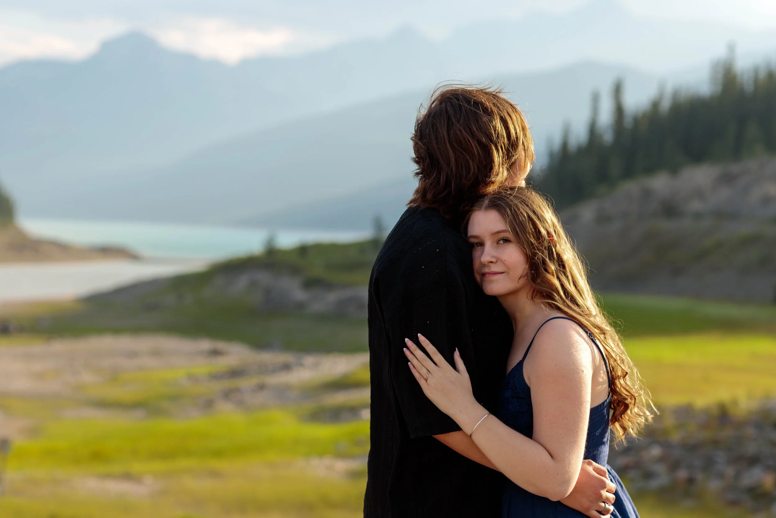 Two people hugging outdoors with mountains and trees in the background, one with red hair and a nose ring, the other with brown hair, both embracing and smiling.