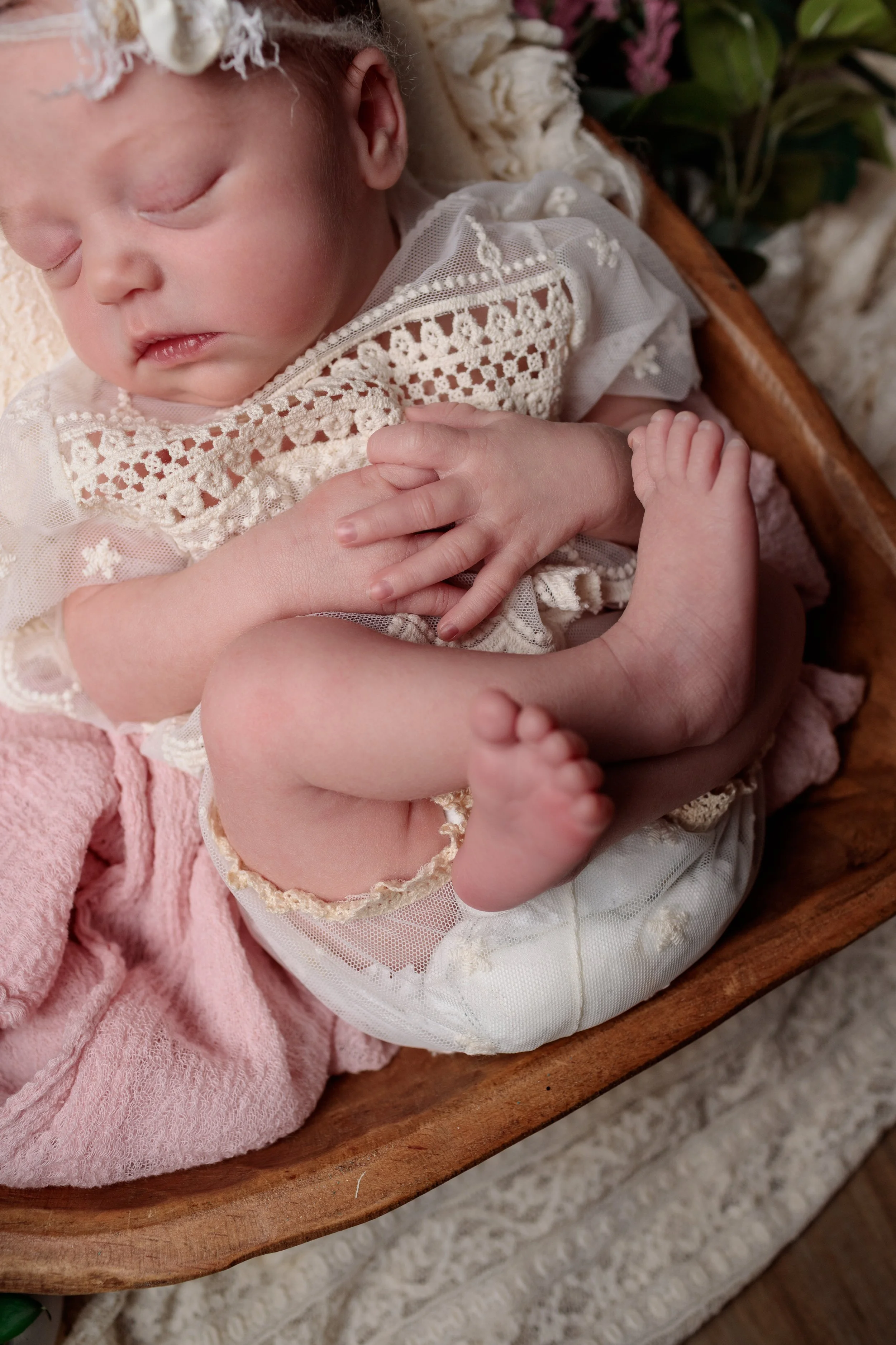 Close-up of a sleeping baby wearing a white lace outfit, holding a tiny baby hand, lying on a pink soft blanket in a wooden cradle. There are some flowers and green leaves in the background.