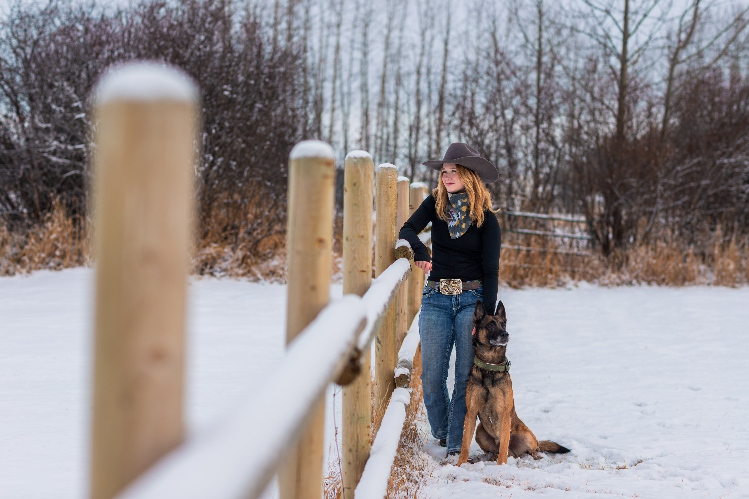 A woman with red hair wearing a wide-brimmed hat, scarf, black shirt, and jeans standing outside in the snow next to a German Shepherd dog, which she is holding with her right hand. They are near a wooden fence with snow on top, surrounded by snow-co