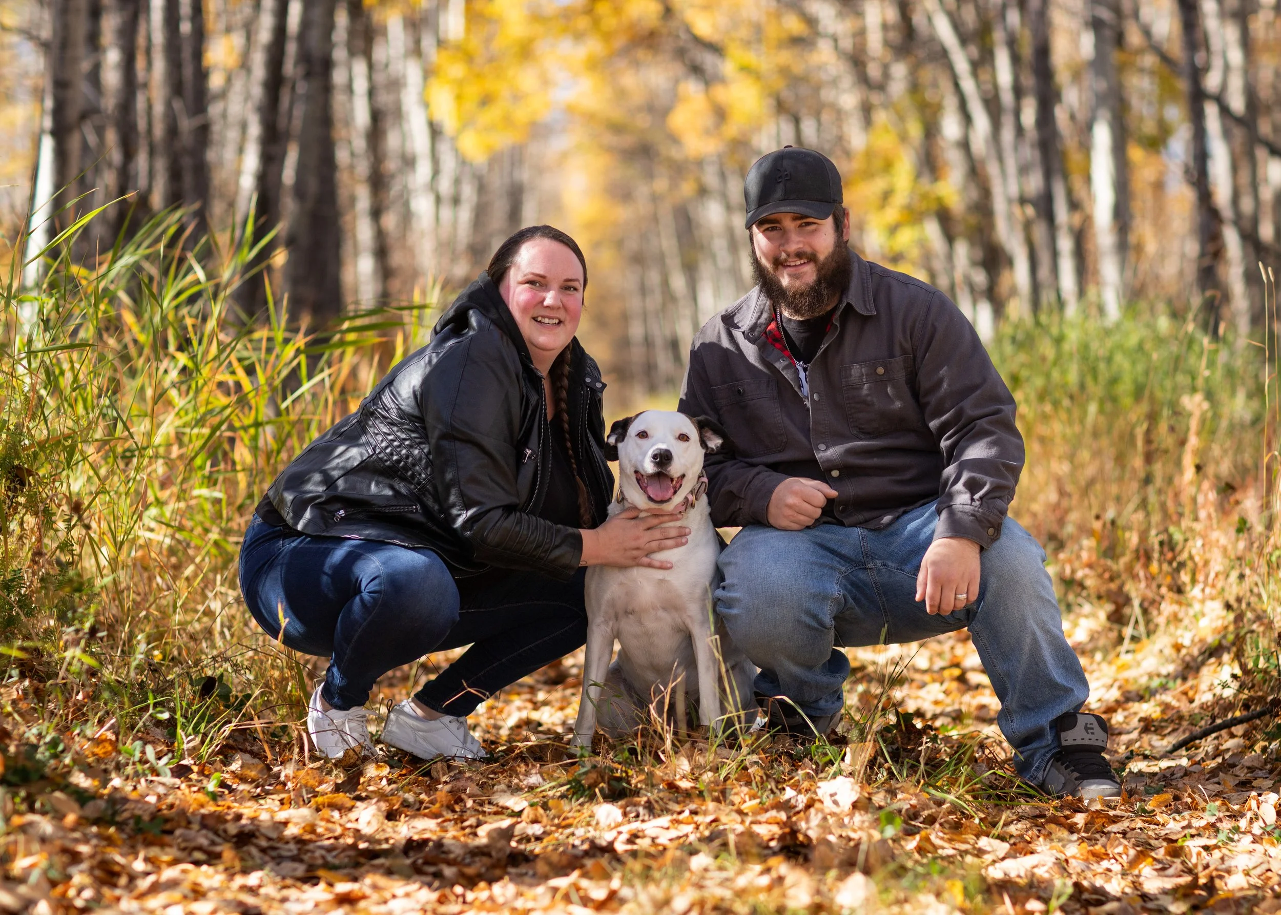 A smiling couple and their dog sitting on the ground in a forest with autumn leaves.