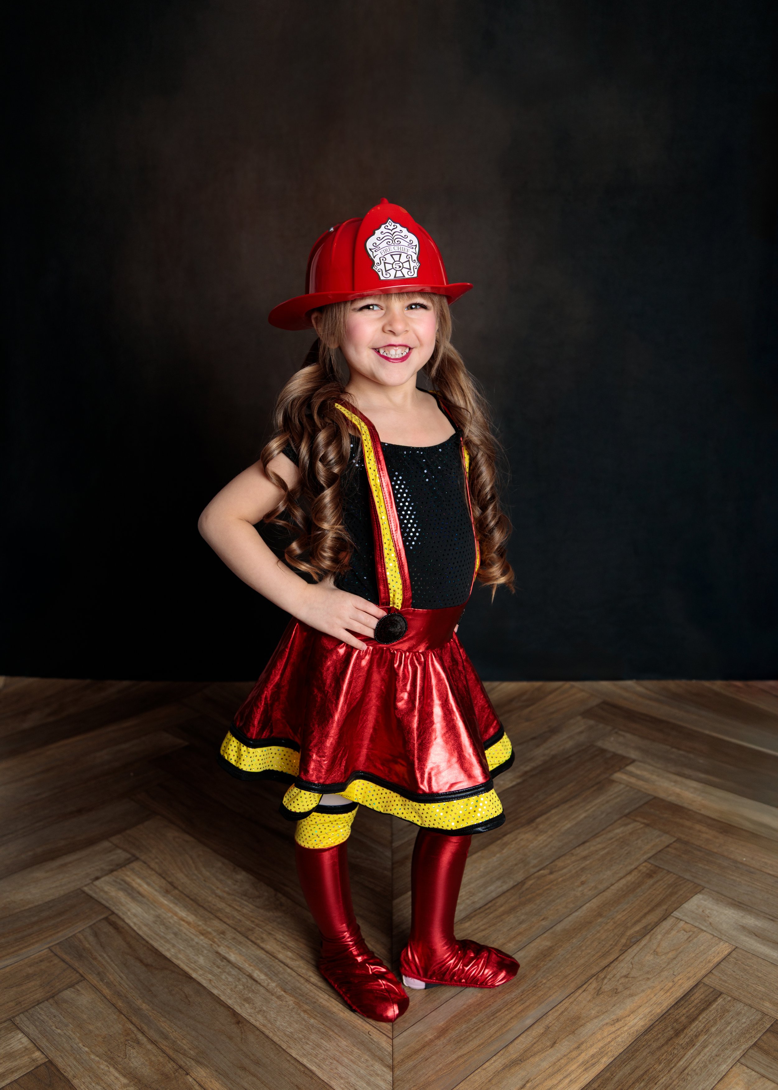 Young girl dressed as a firefighter with a red fireman's hat, a shiny black top, a red skirt with yellow and black trim, yellow polka dot leggings, and red shiny boots, standing with her hand on her hip and smiling.