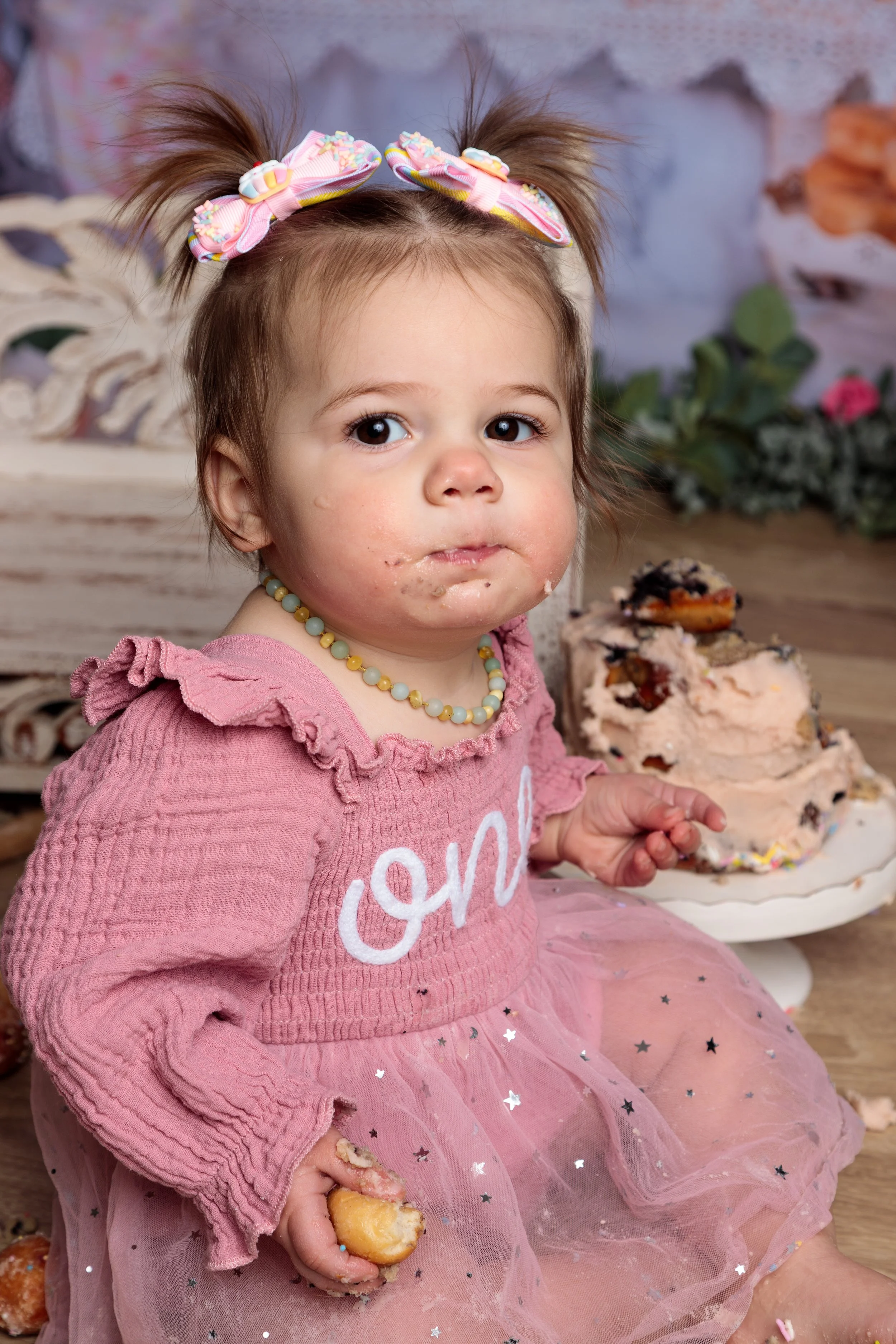 A young girl with brown hair tied in two pigtails with pink bows, wearing a pink top, pearl necklace, and a pink tutu skirt, eating a slice of cake with frosting and sprinkles, with some frosting on her face and hands, sitting in front of a plate wit