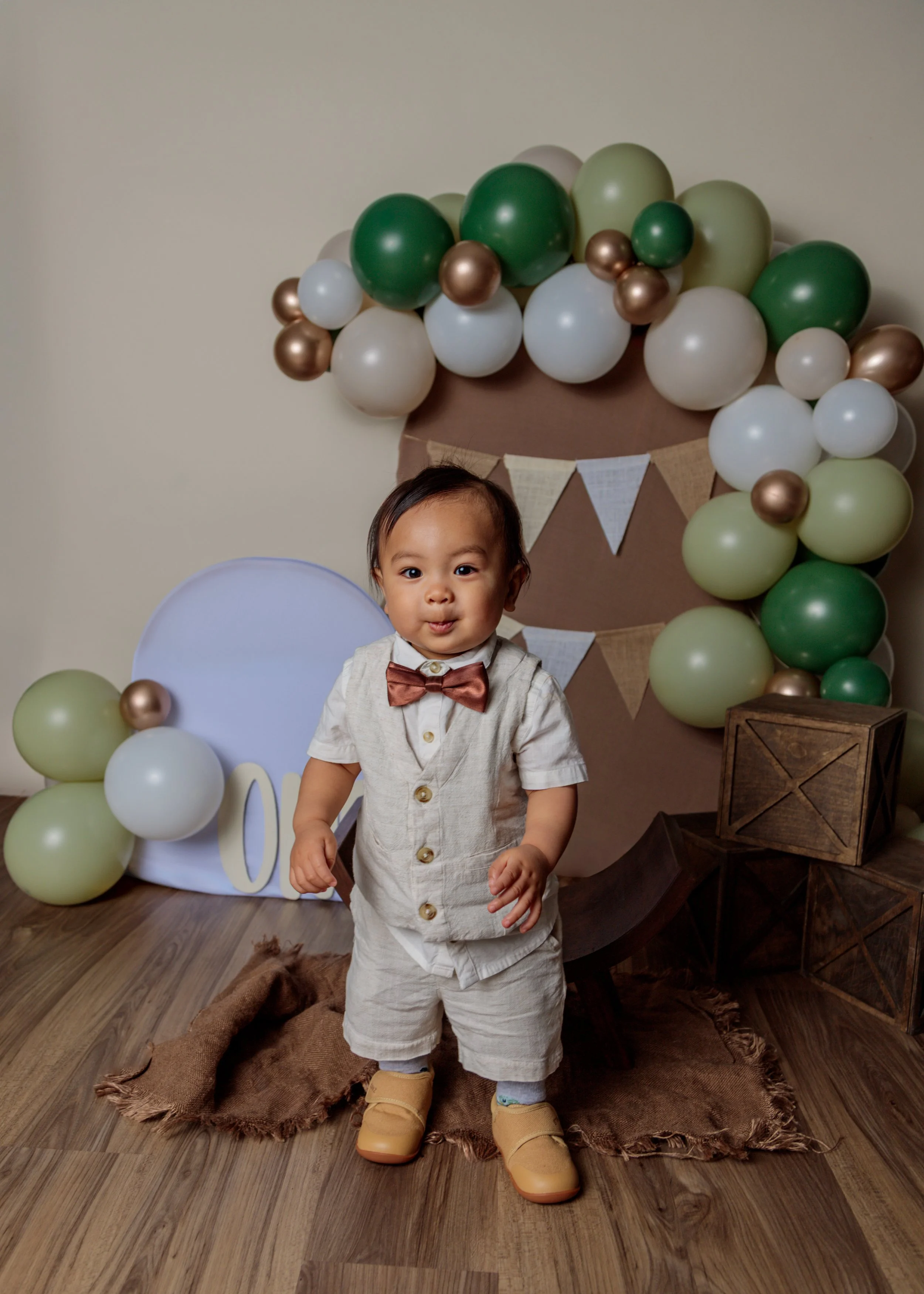 A young boy dressed in a cream-colored outfit with a bow tie and tan shoes, standing on a wooden floor in front of a balloon backdrop with green, white, and gold balloons, and decorative banners. His hands are slightly raised and he's looking at the 