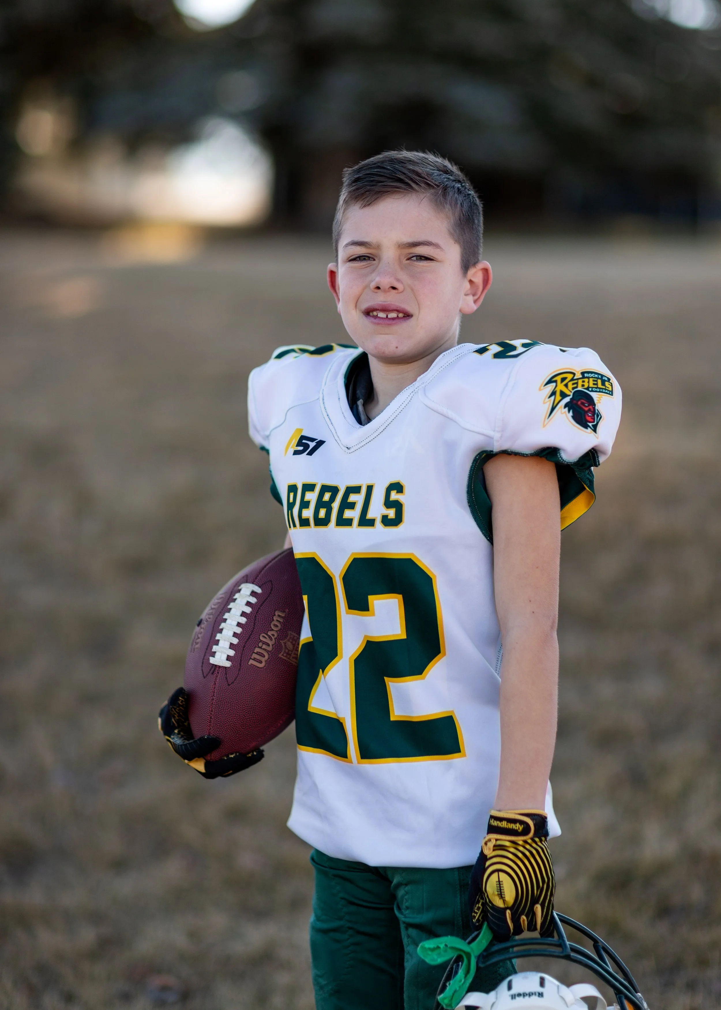 Young boy wearing a Rock Jets Rebels football jersey, holding a football, and standing outdoors.