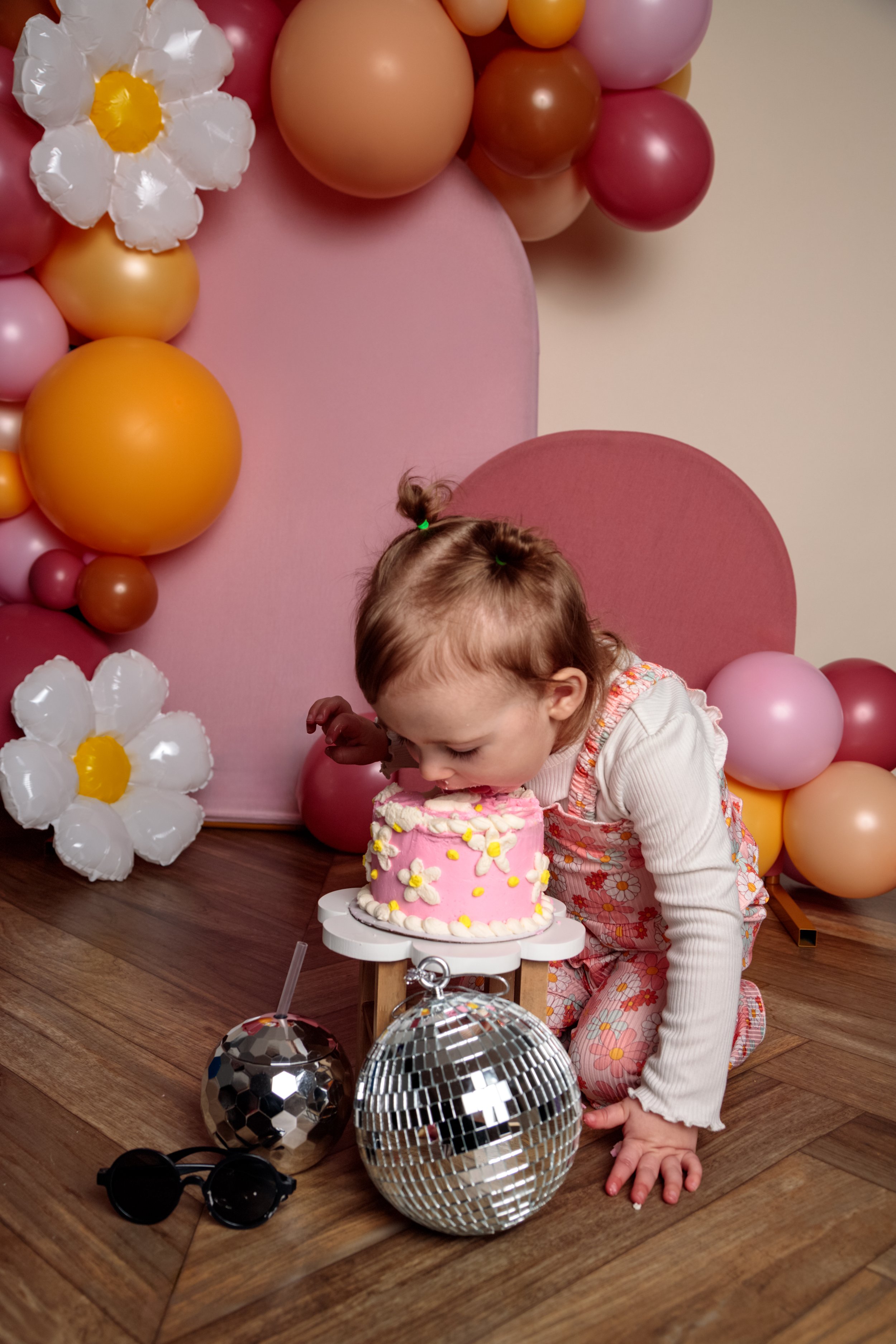 A young girl wearing a floral dress is celebrating her birthday, sitting next to a pink birthday cake with white and yellow decorations. The background features a pink and beige balloon arch and white and yellow paper flowers. Two disco balls are on 