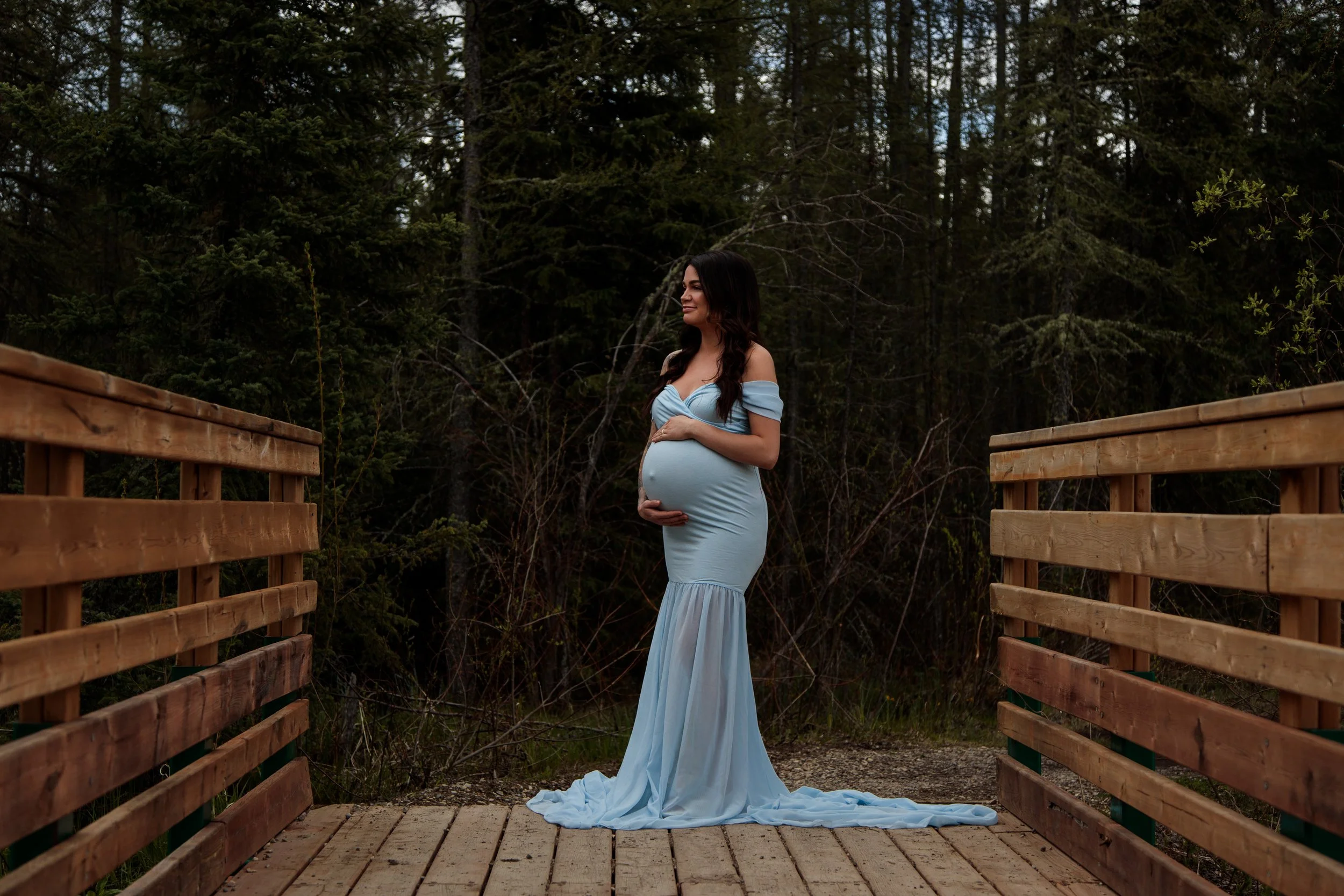 A pregnant woman in a flowing light blue dress standing on a wooden bridge in a forest, holding her belly and smiling.