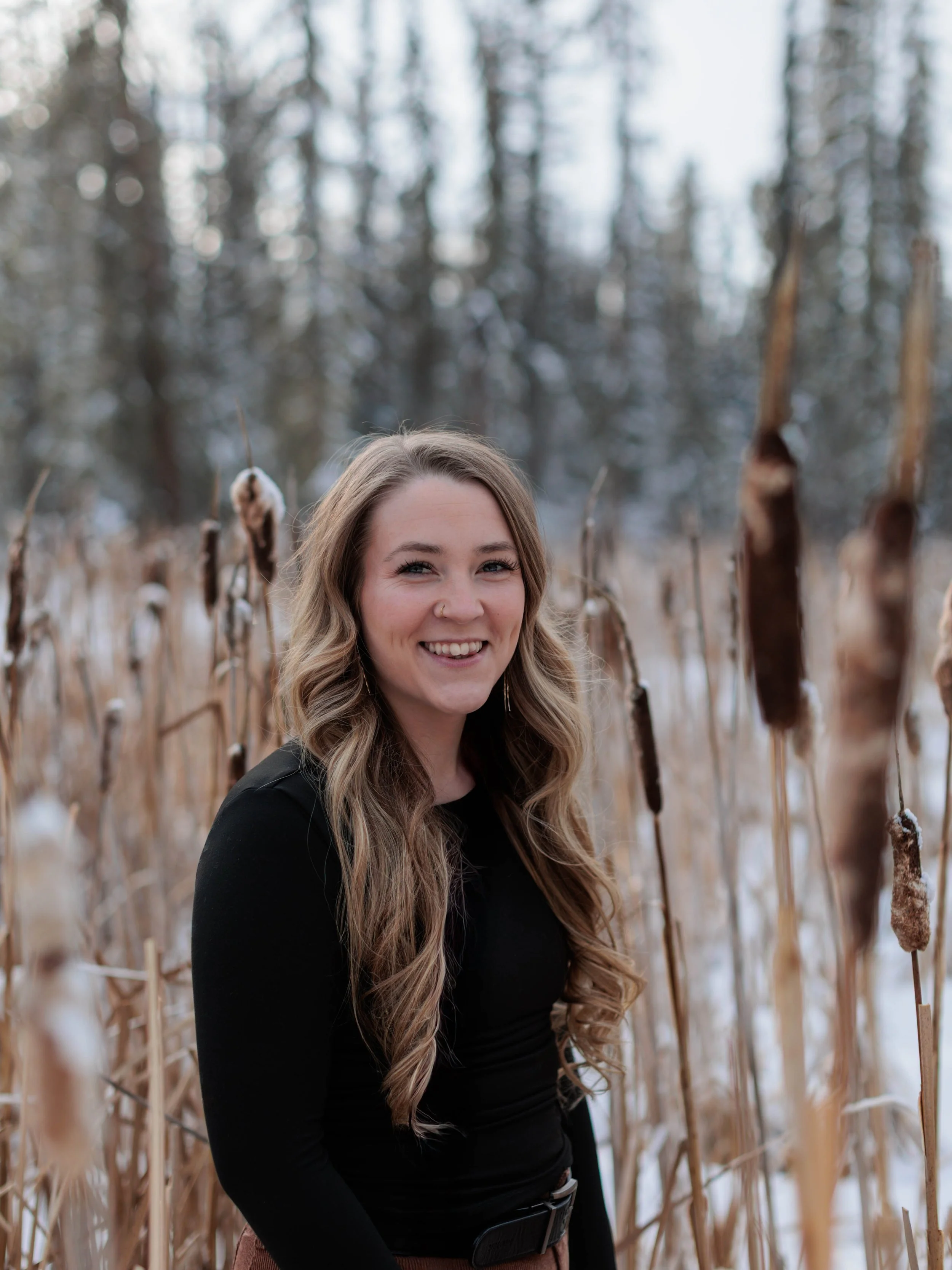 A woman with long, wavy blonde hair, smiling, standing in front of dry, brown cattails in a winter field, with a blurred background of trees.