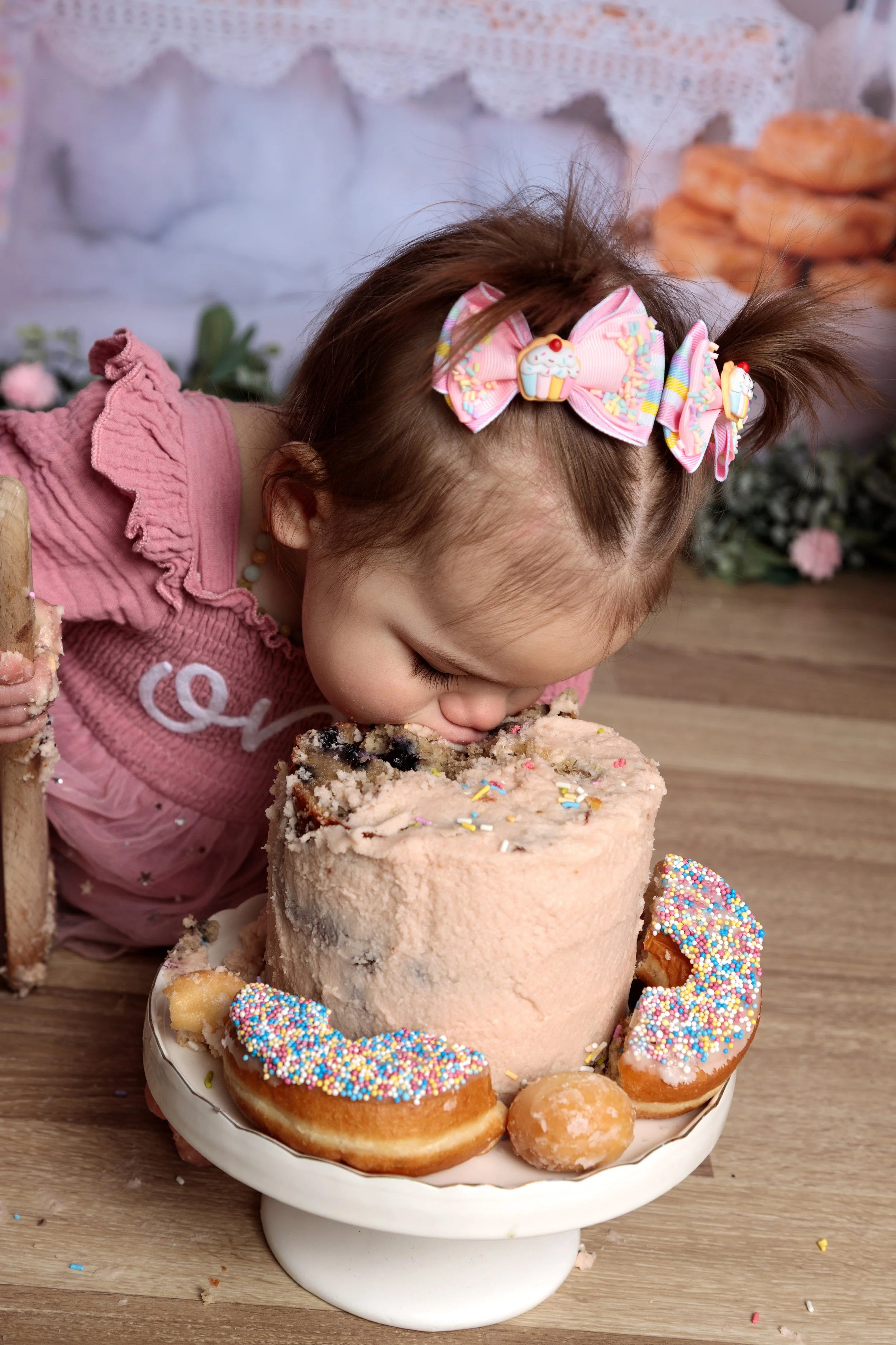 Young girl with a pink bow hair clip decorated with cupcakes and sprinkles, leaning over a console with a large cake topped with pink icing, colorful sprinkles, and surrounded by decorated donuts and cookies with rainbow sprinkles.
