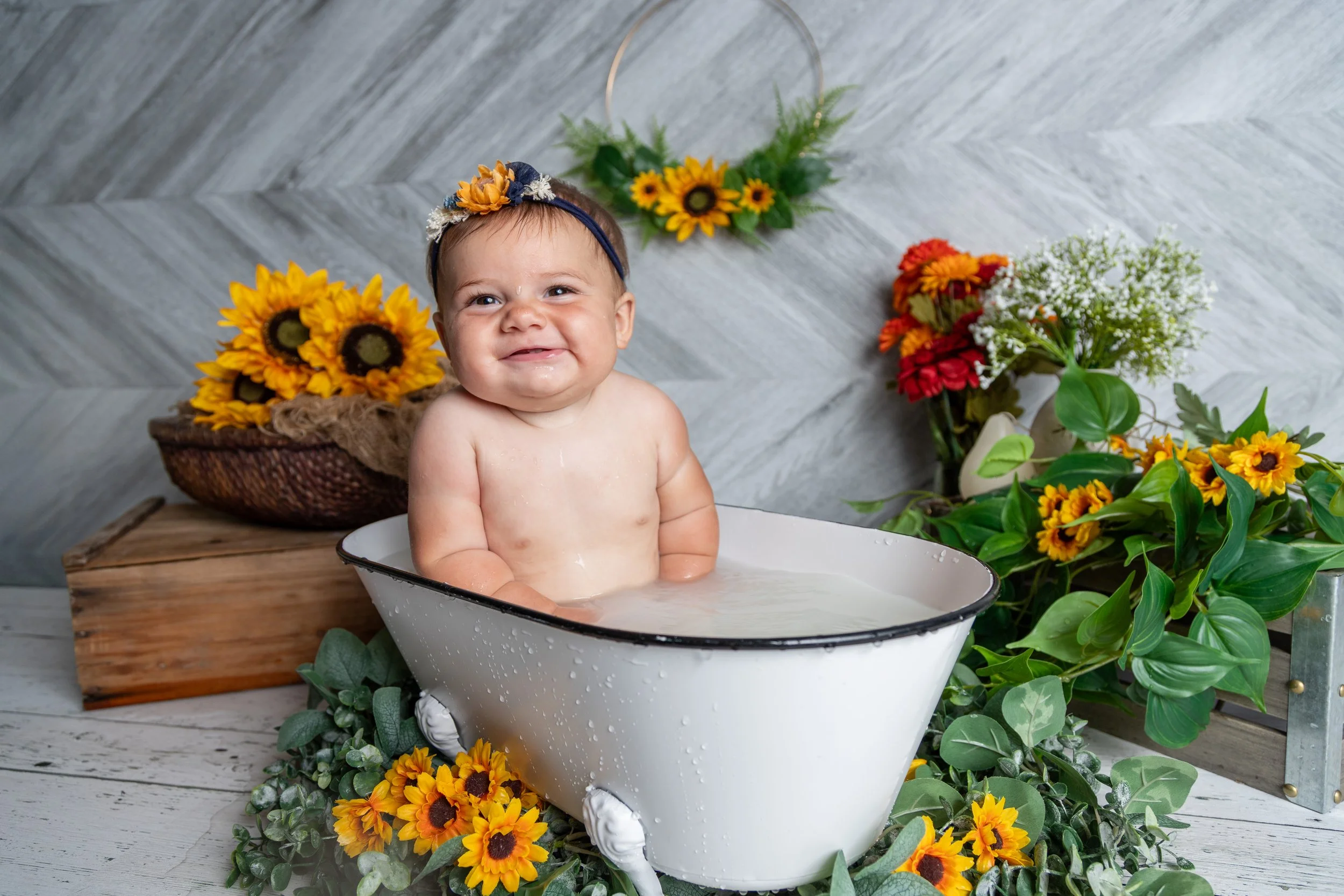 A smiling baby with a floral headband taking a bath in a white enamel tub, surrounded by yellow, red, and white flowers and green foliage, with a grey wooden floor and grey textured wall background.