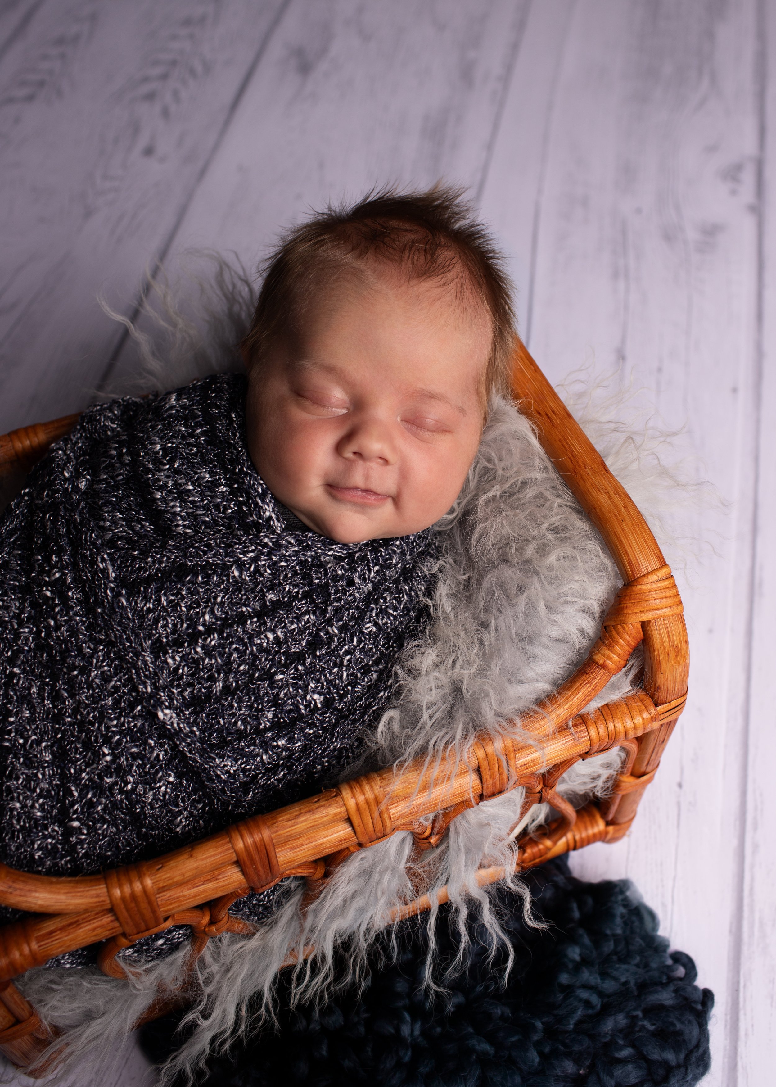 A baby is sleeping in a wicker basket lined with soft fur, on a white wooden surface.