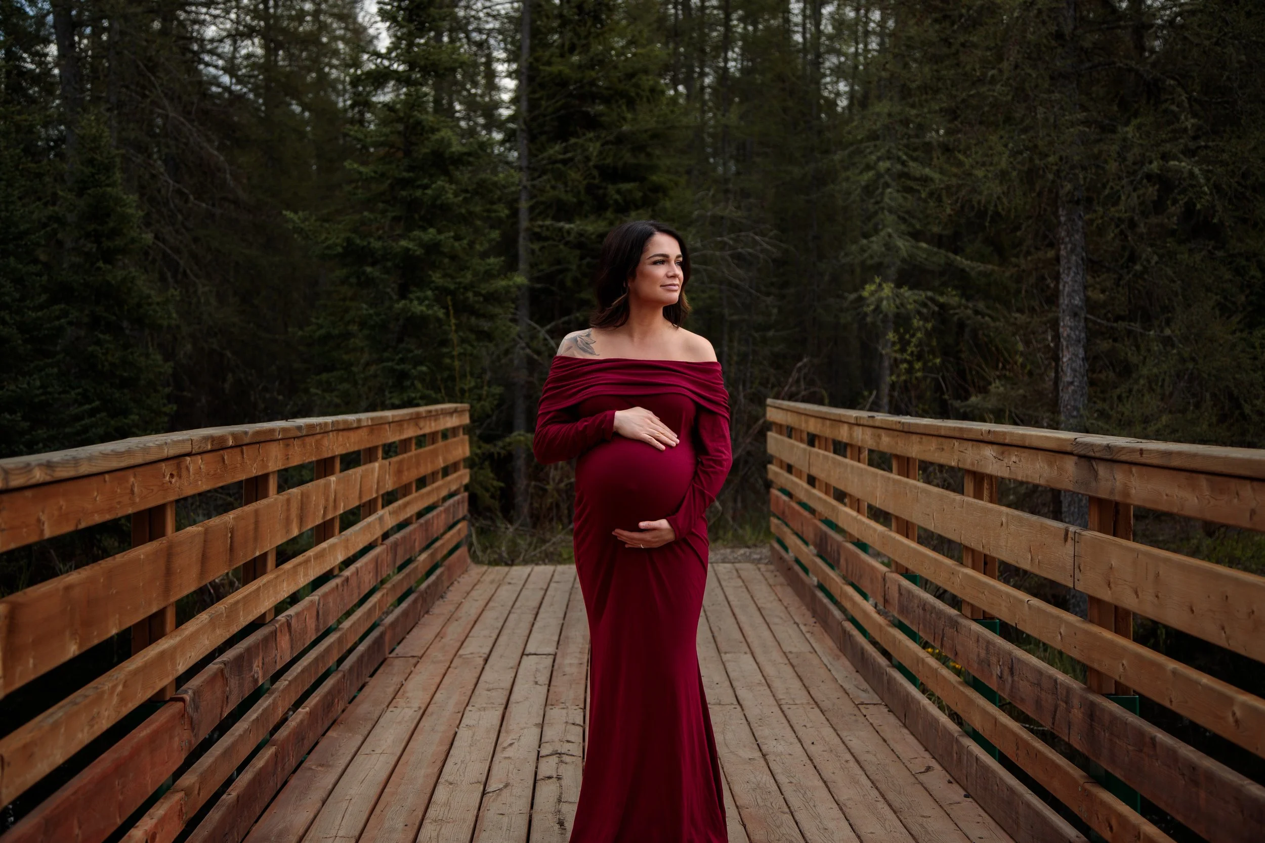 A pregnant woman wearing a long red off-the-shoulder dress standing on a wooden bridge with a forest background.
