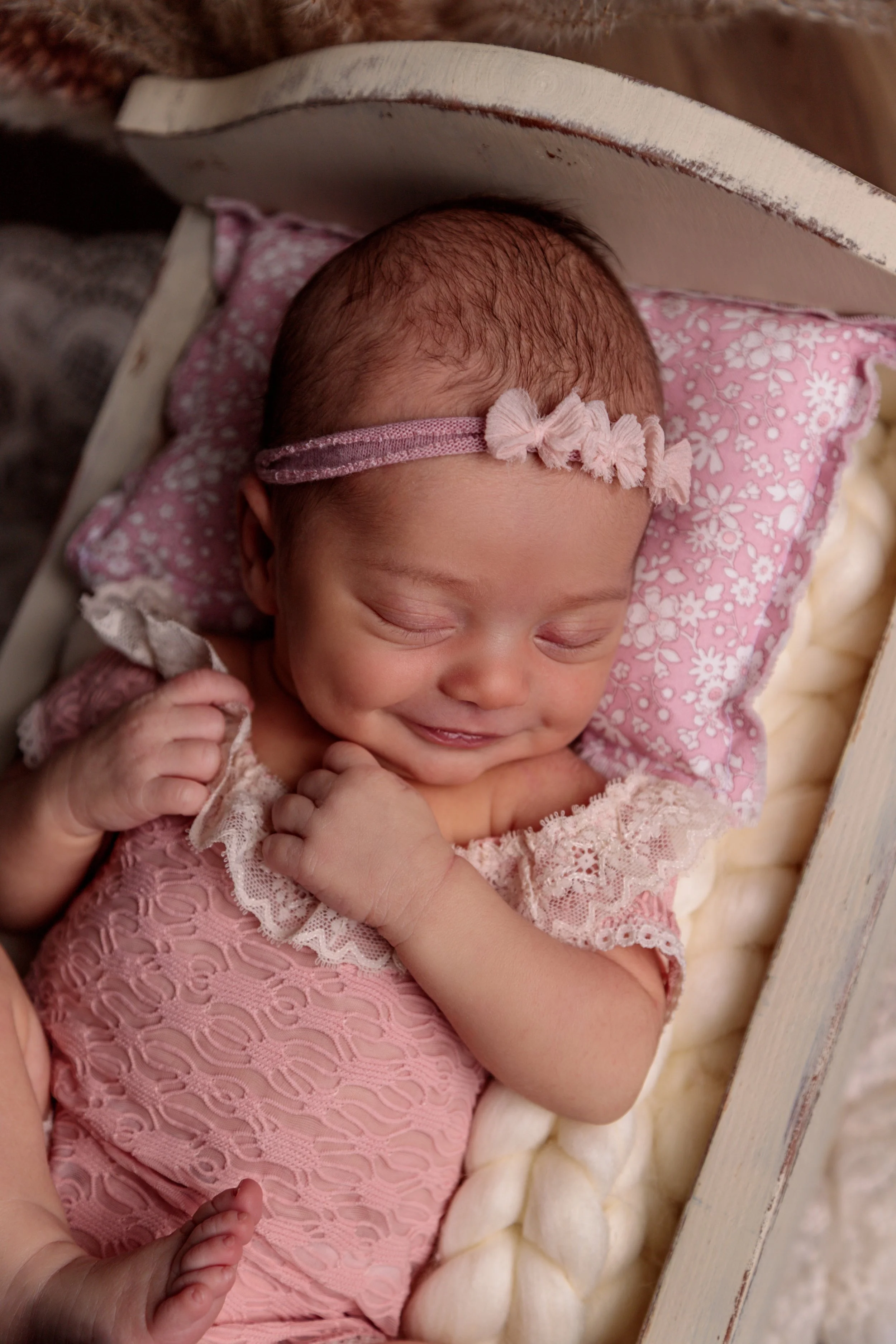 A sleeping baby girl with a pink headband with flowers, dressed in a pink lace dress, lying on a pink floral pillow inside a wooden cradle, with a soft cream-colored blanket underneath.