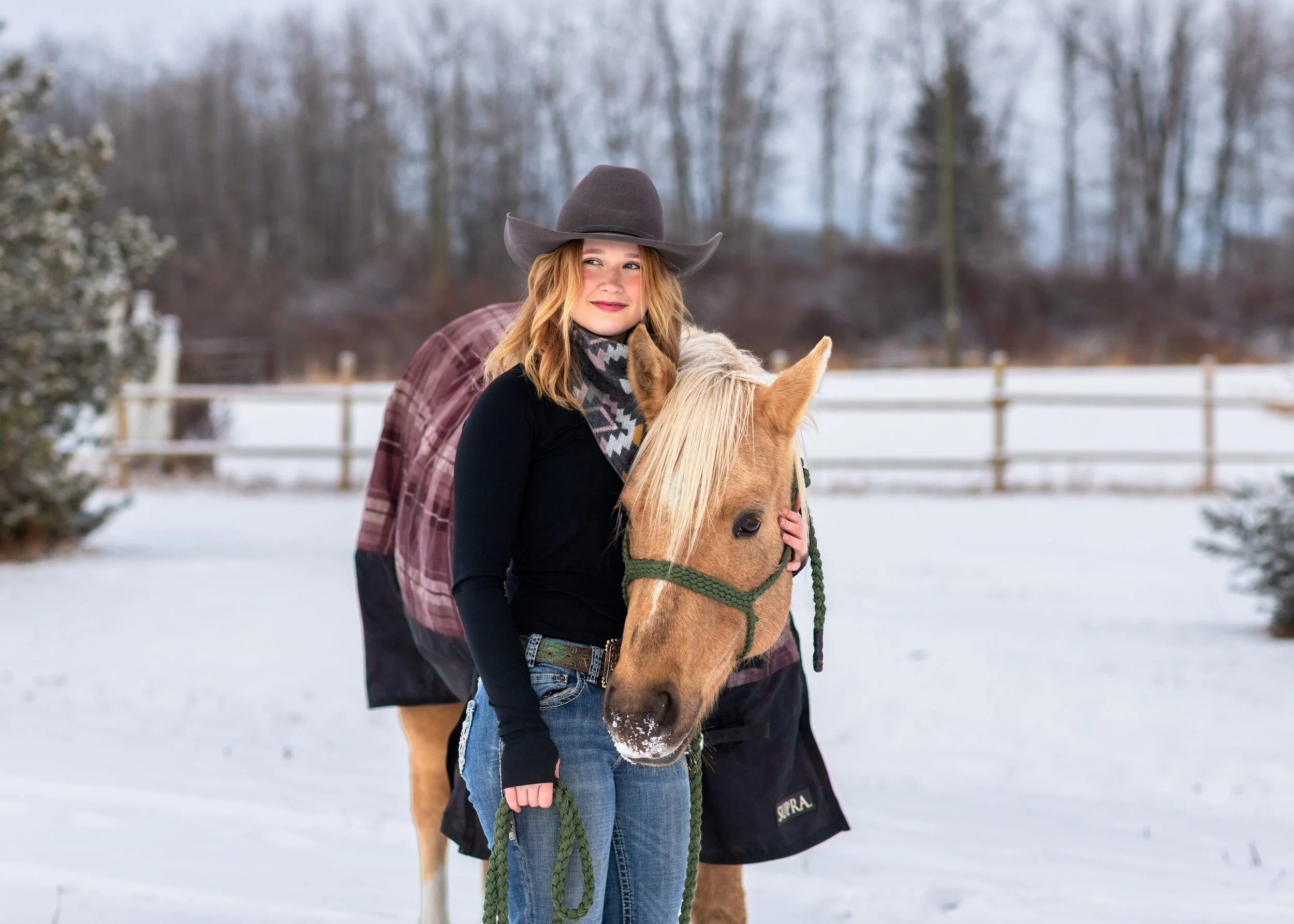 A woman with red hair wearing a gray cowboy hat and a black shirt stands next to a young girl on a light brown horse with a green harness in a snowy outdoor area. A man in a plaid shirt stands behind them, holding the horse's saddle. The background s