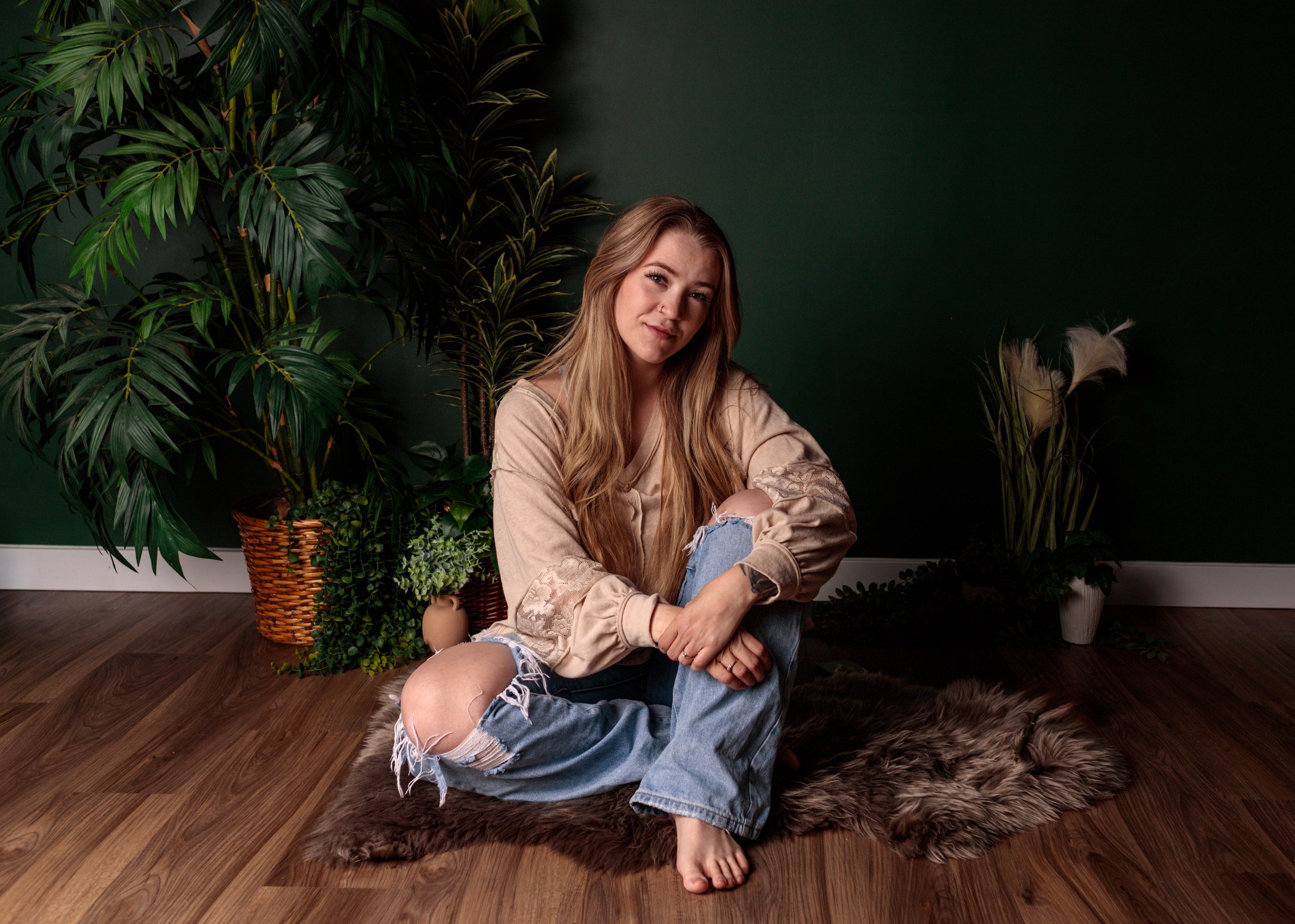 A young woman with long blonde hair sitting on the floor amidst large green houseplants, with a dark green wall in the background.