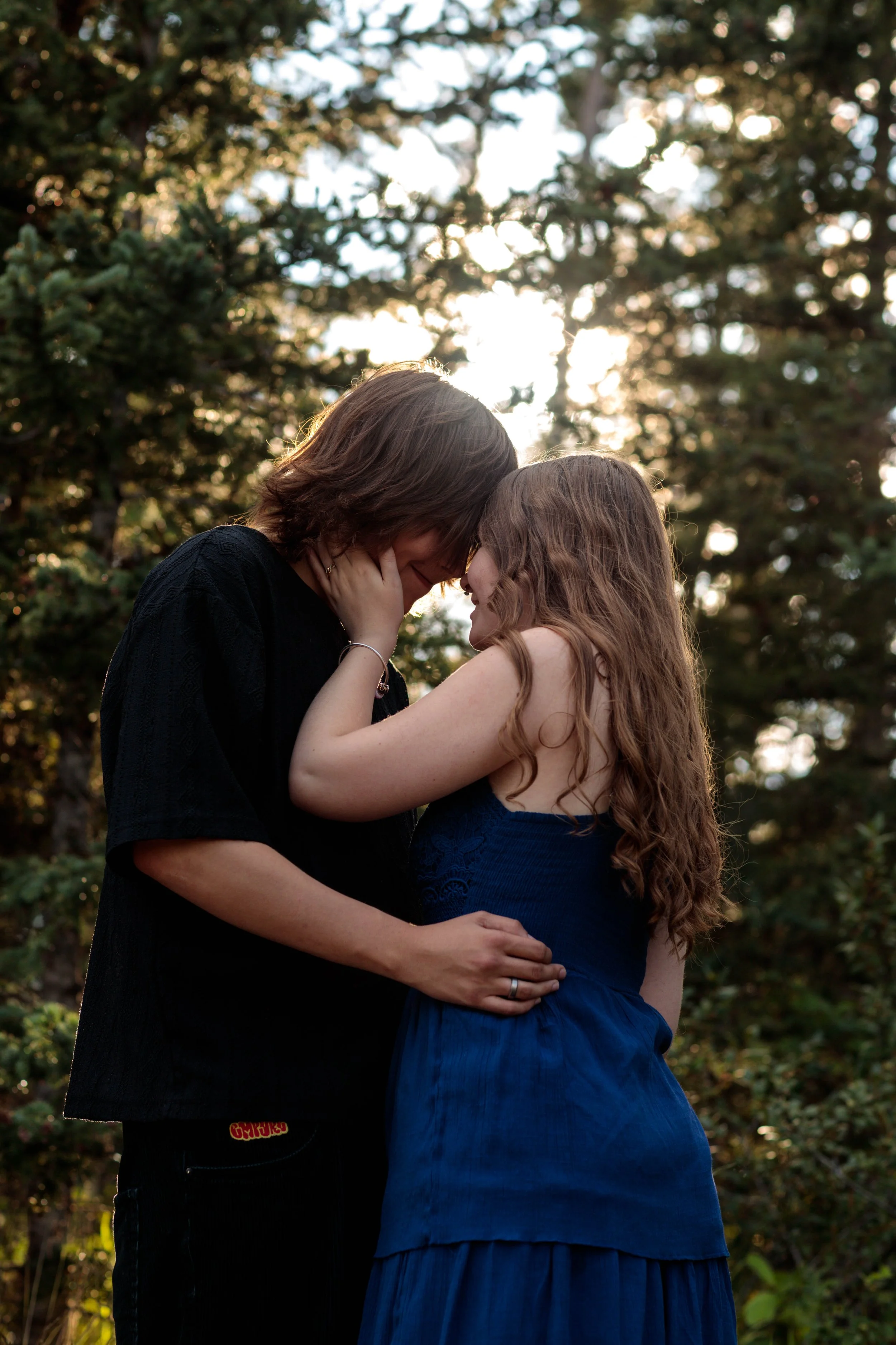 A couple sharing an intimate moment outdoors during sunset. The man and woman are touching foreheads, with the man's hands around the woman's waist. The background shows trees with sunlight shining through.